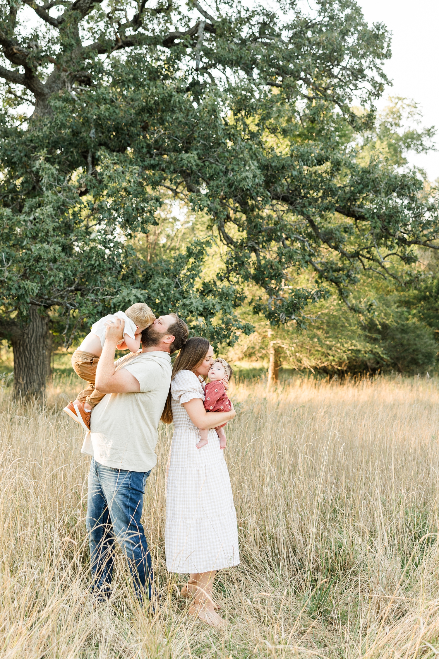 The Mathisens stand back to back in a grassy pasture while holding up both of their small children for kisses | CB Studio