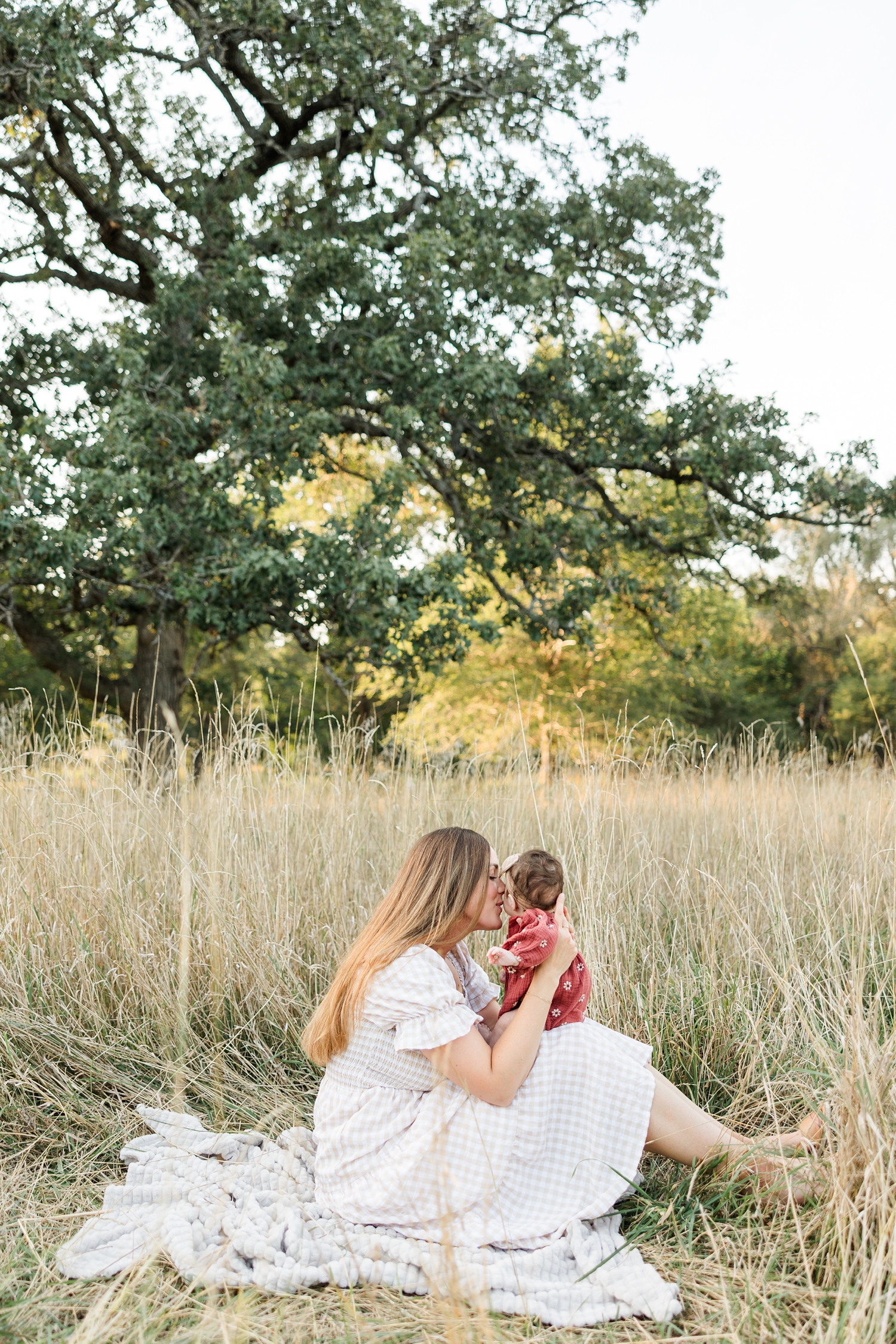 Allyson sits in a grassy pasture surrounded by tall grasses and oak trees while kissing her new baby girl | CB Studio