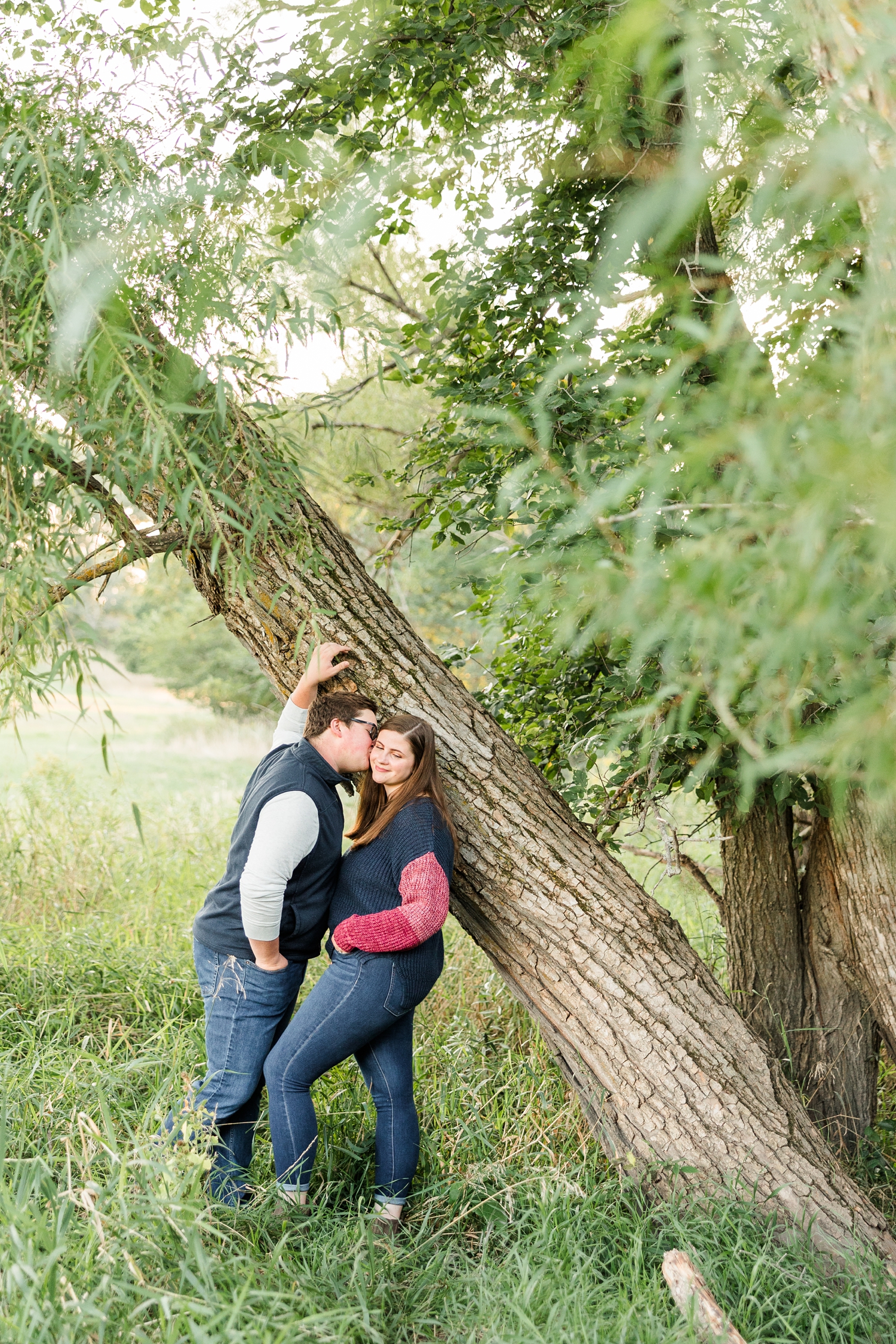 Kevin kisses Myra's cheek as they lean against a tree surrounded by light green leaves and branches | CB Studio