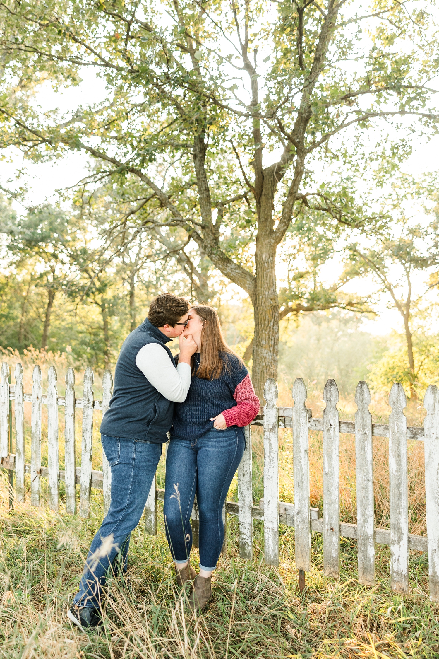 Kevin and Myra, while leaning on a rustic, white picket fence, share a kiss in the middle of a grass pasture surrounded by trees | CB Studio