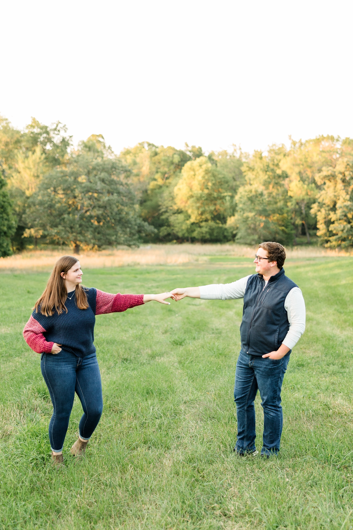 Myra and Kevin hold hands as far apart as they can in the middle of a grassy pasture with a beautiful tree line in the distance | CB Studio