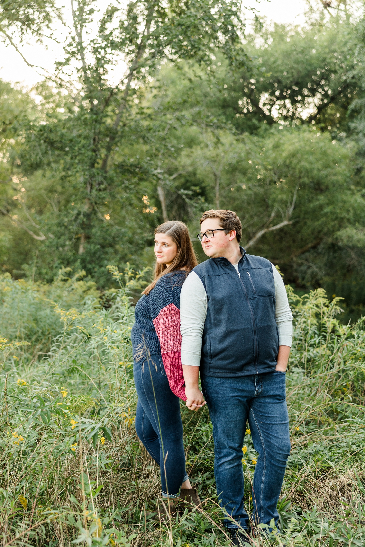 Kevin and Mrya stand with their backs towards each other, leaning on each other's shoulders and holding hands as they look off to the left in the distance in the middle of a tree filled grassy pasture | CB Studio