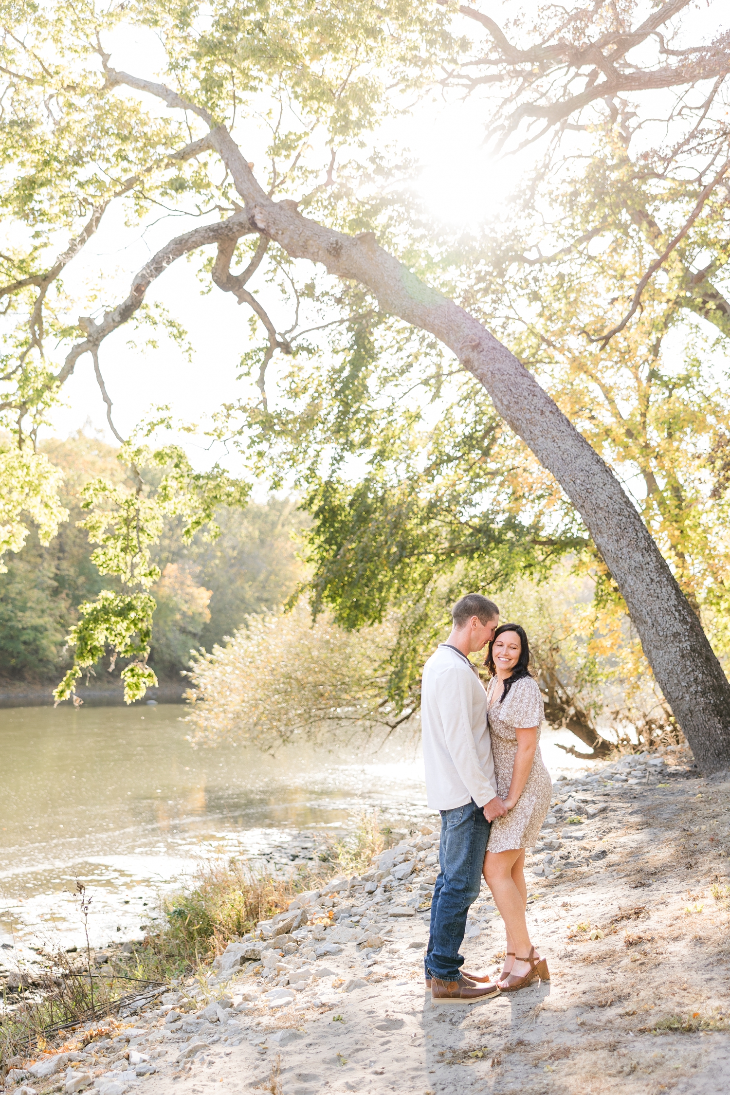 Brandon and Haley stand together holding hands on a sandy, Cedar River beach with a curved tree in the background as Brandon nuzzles Haley gently, located in Osage IA | CB Studio