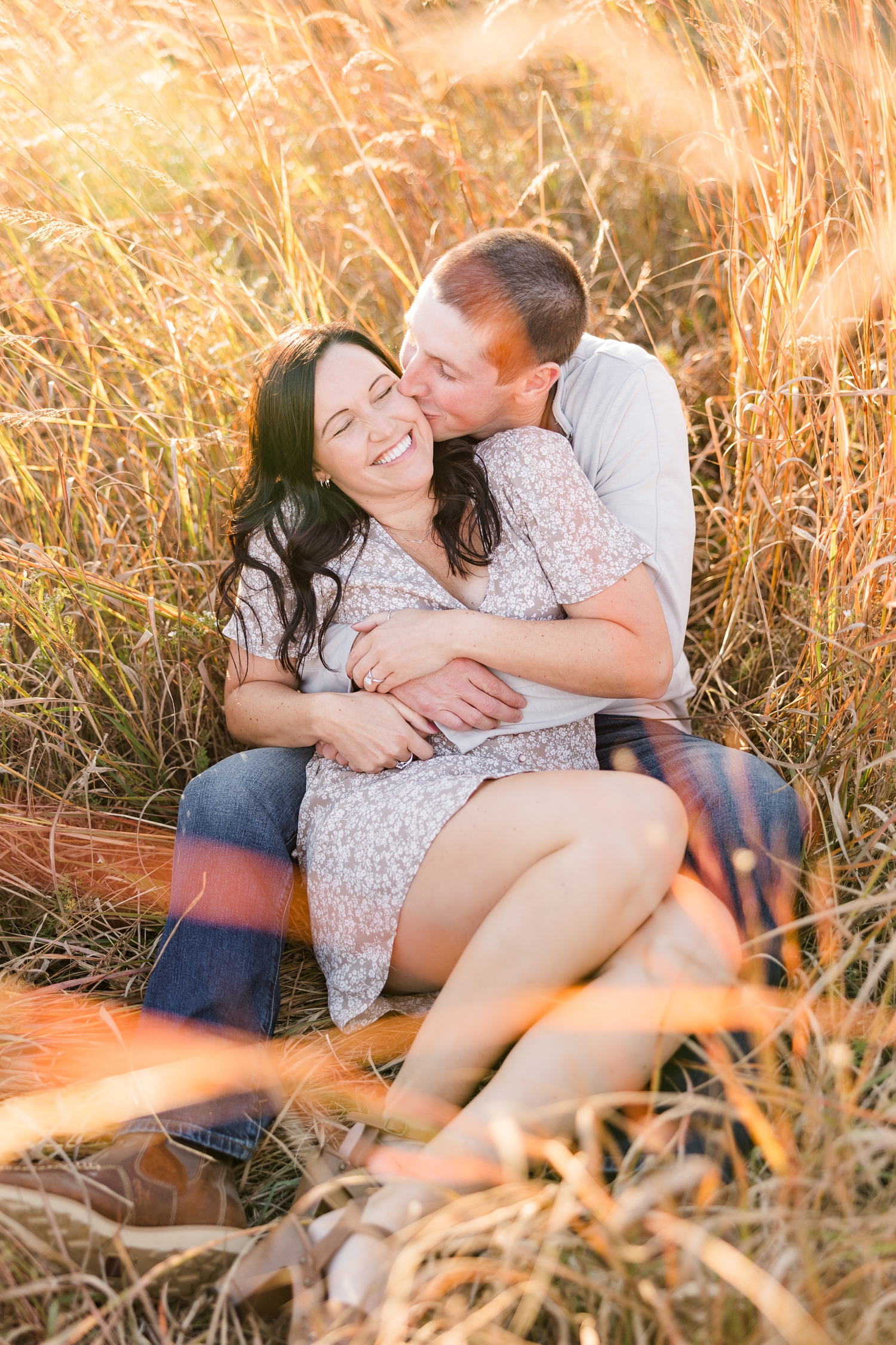 Brandon and Haley sit together in a grass field surrounded by orange fall grass as Brandon squeezes Haley's waist and pulls her in for a kiss on the cheek, located in Osage IA | CB Studio