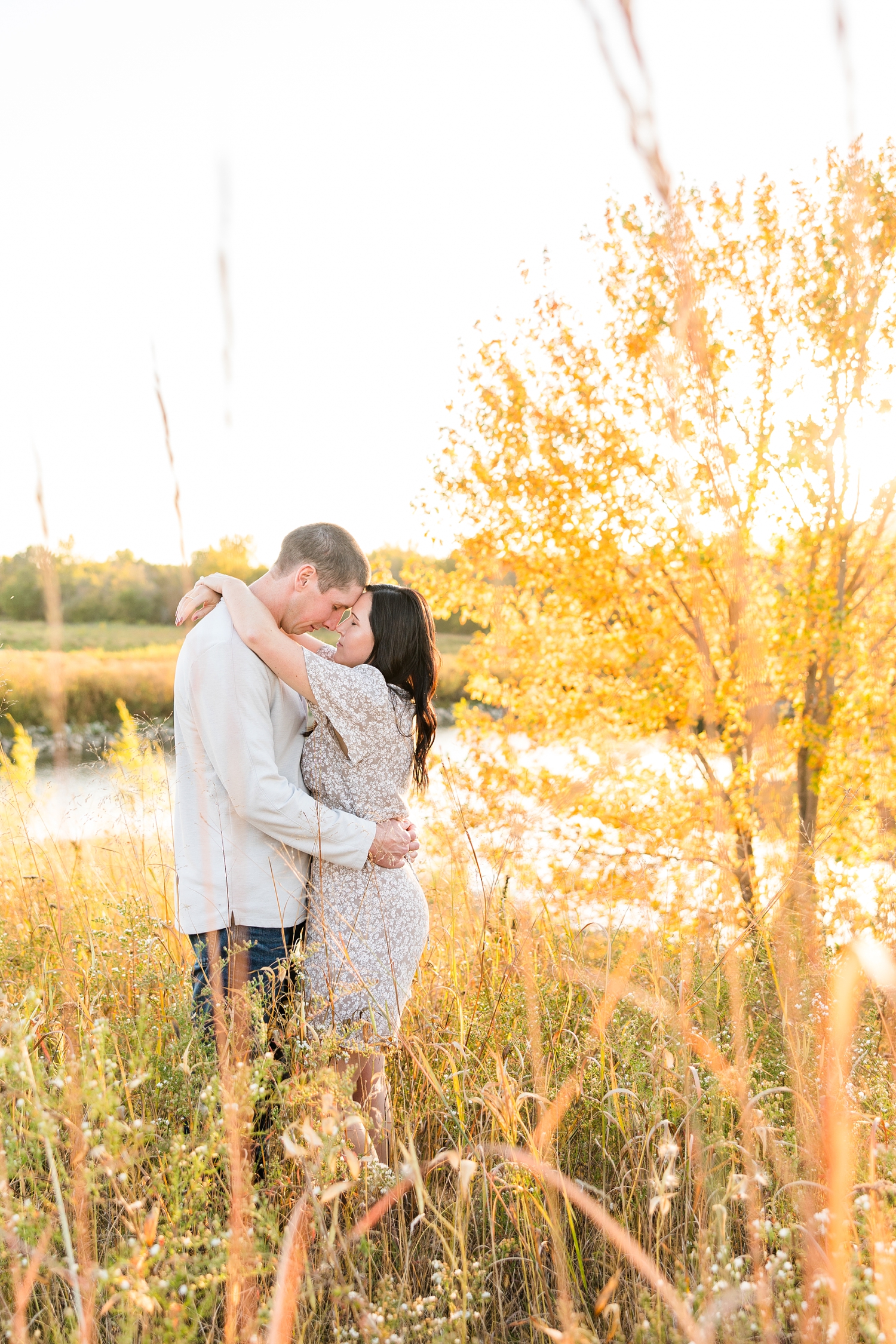 Brandon and Haley embrace with Haley's arm around Brandon's shoulders and their foreheads resting together as they stand in a fall grass field with a tree full of orange/yellow foliage and a pond in the background, located in Osage IA | CB Studio