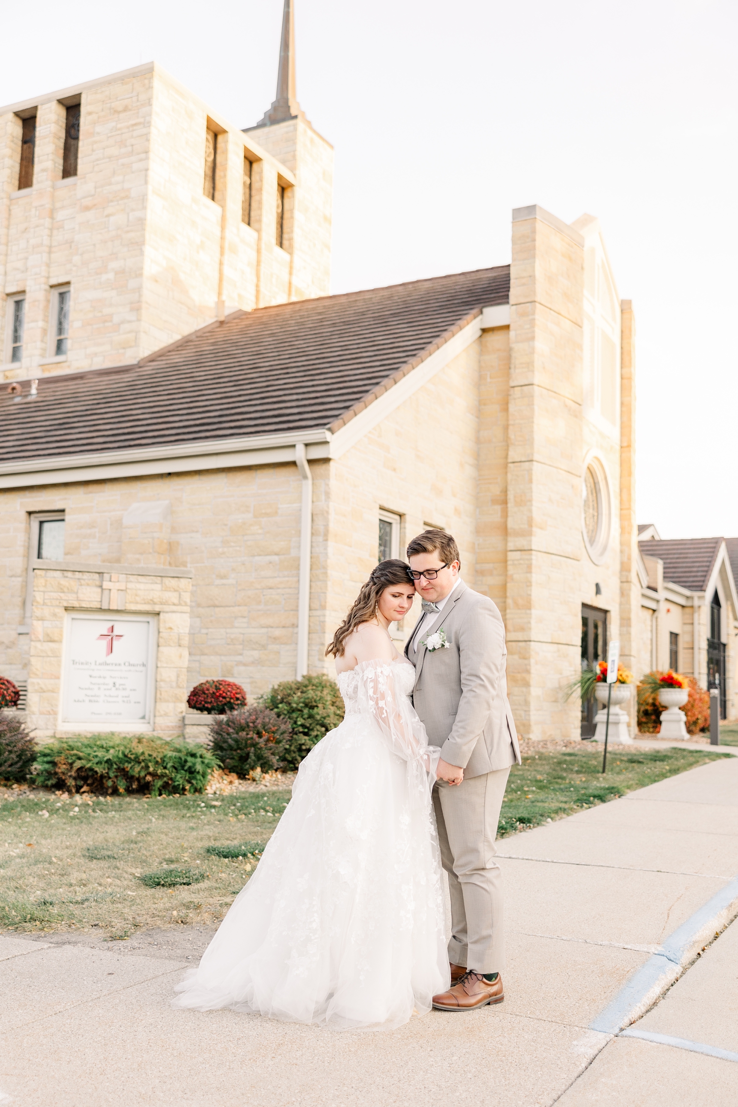 Kevin and Myra stand together holding hands and looking in front of Trinity Lutheran Church in Algona, IA during sunset on their wedding day | CB Studio