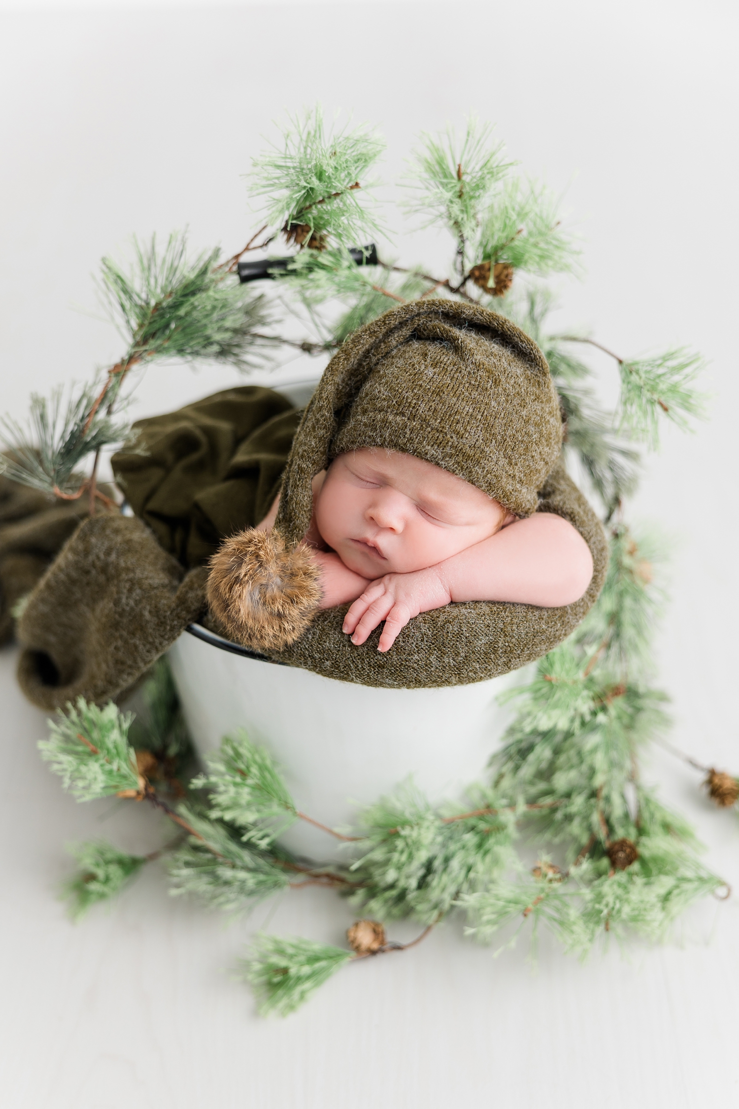 Baby Joey, wearing a green wool night cap, nestled in a white bucket surrounded by an evergreen garland | CB Studio