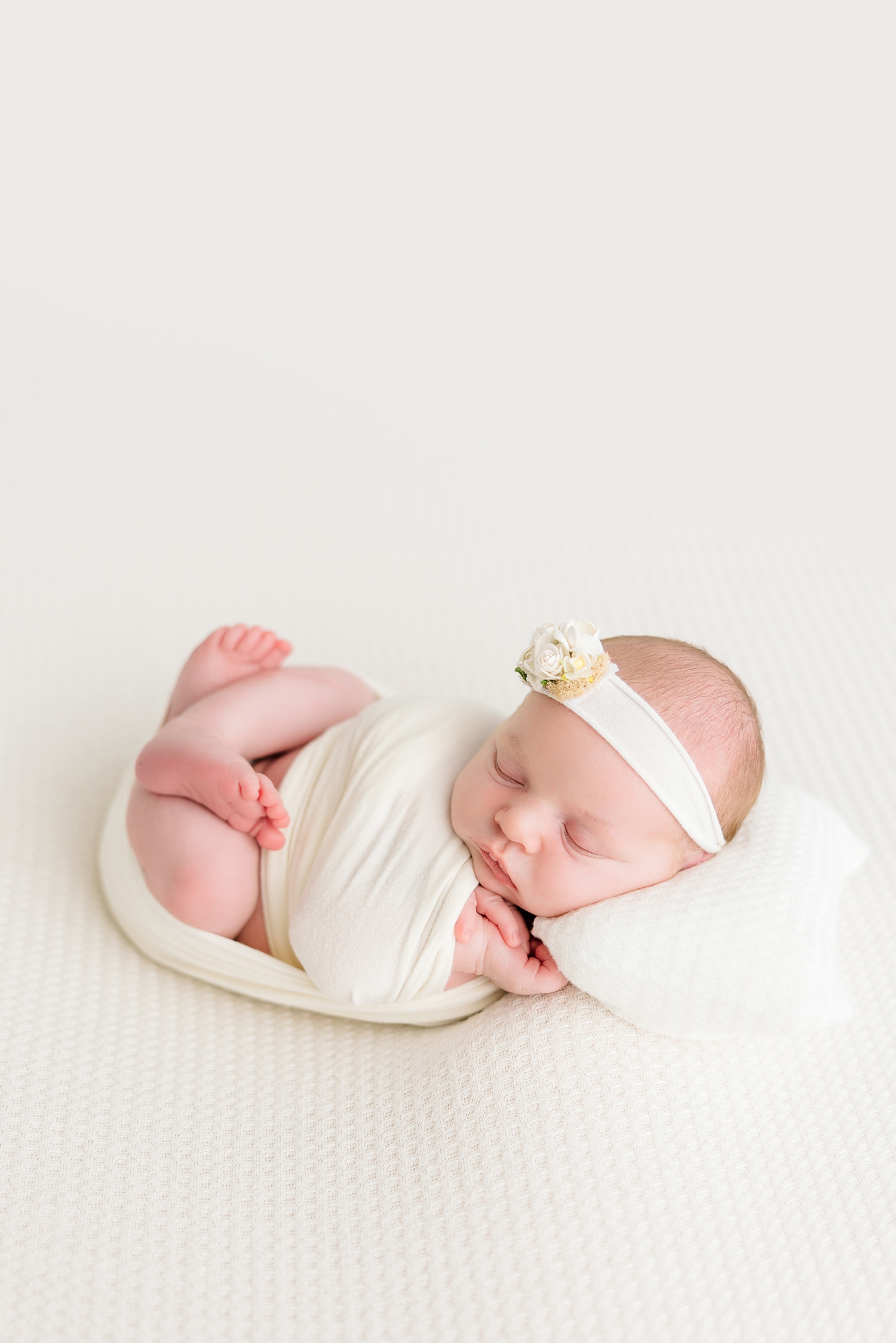 Baby Sadie wrapped and surrounded in all white wearing a white floral headband and resting her head on a white pillow | CB Studio