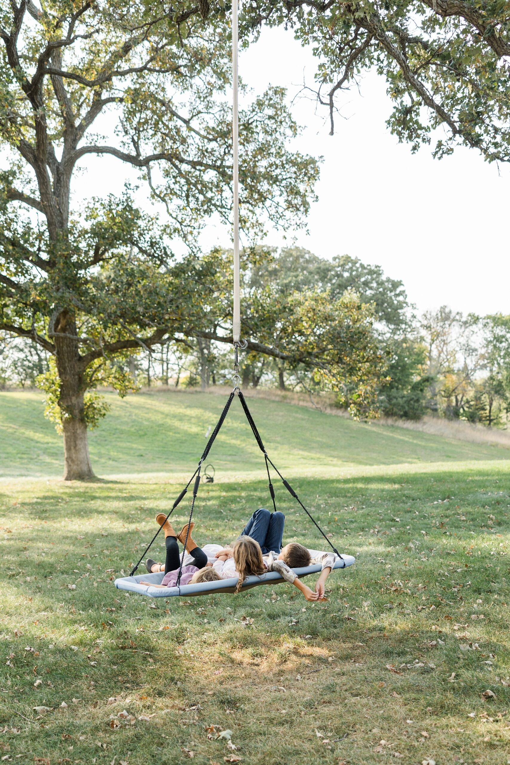 Best of 2024 Families | Three children lay on a large yard swing as they watch the autumn leaves fall | CB Studio