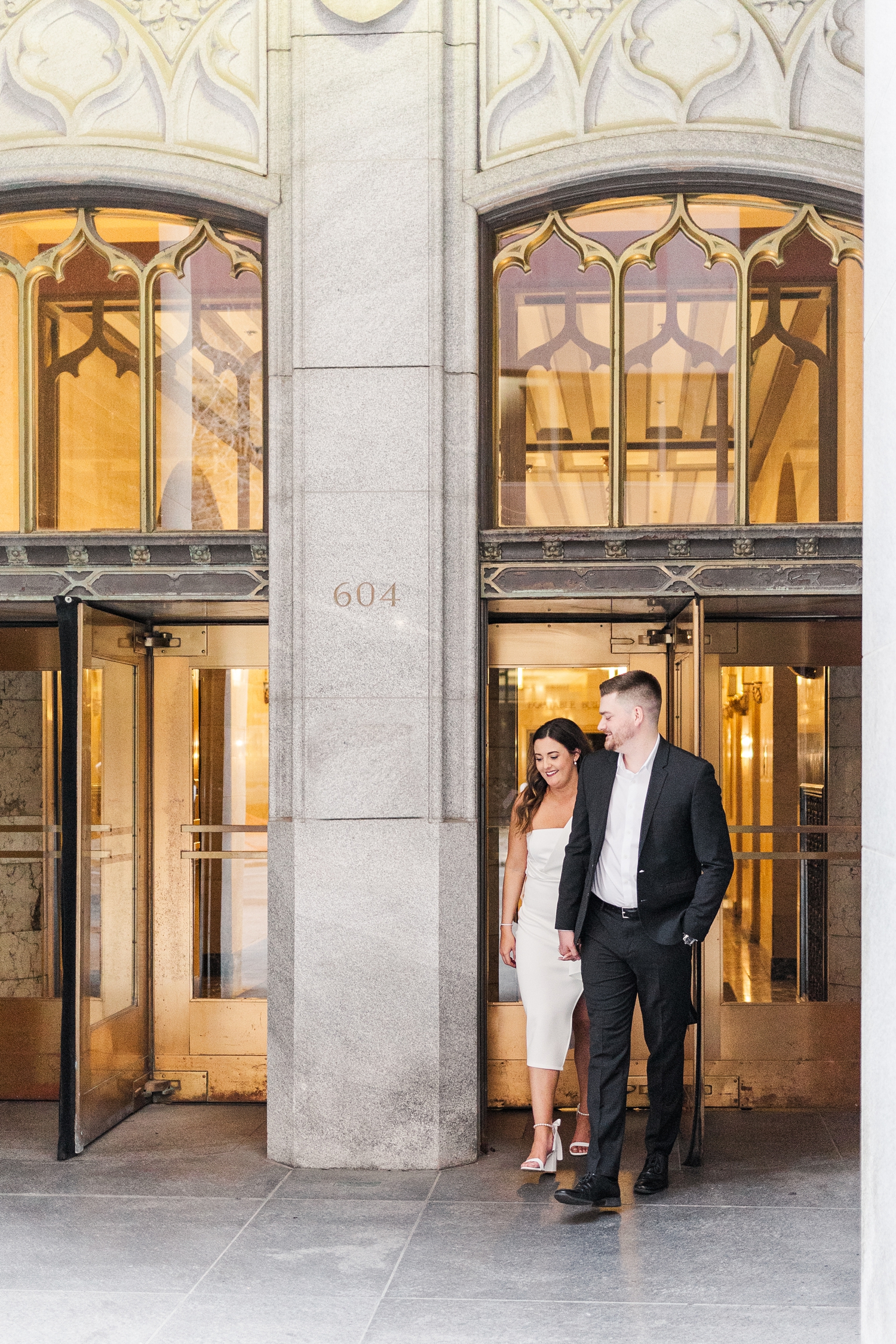 Best of 2024 Engagements | Jenna and Dustin walk hand in hand as they walk out of the intricate gold doors of the The Equitable Building in Des Moines, IA | CB Studio