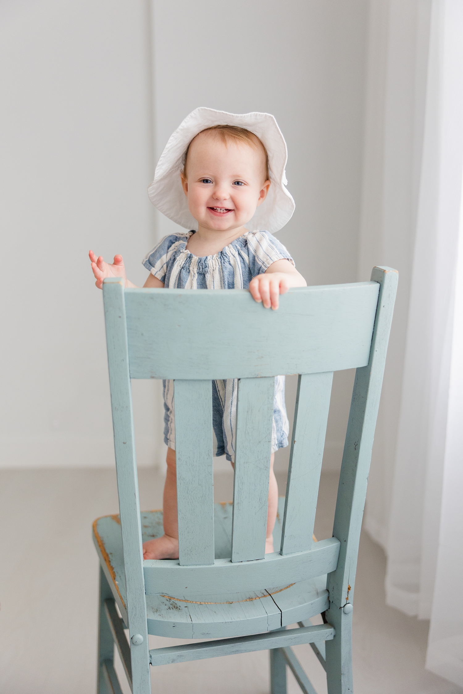 Baby Elnora smiles while standing on a blue chair while holding onto the back of it wearing a white beach hat | CB Studio