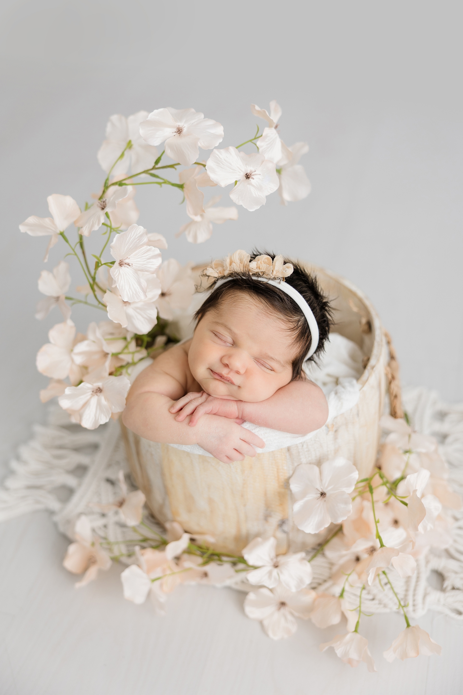 Baby Lennon smiles as she's nestled in a light wood bucket surrounded by apple blossoms in a white wood room | CB Studio