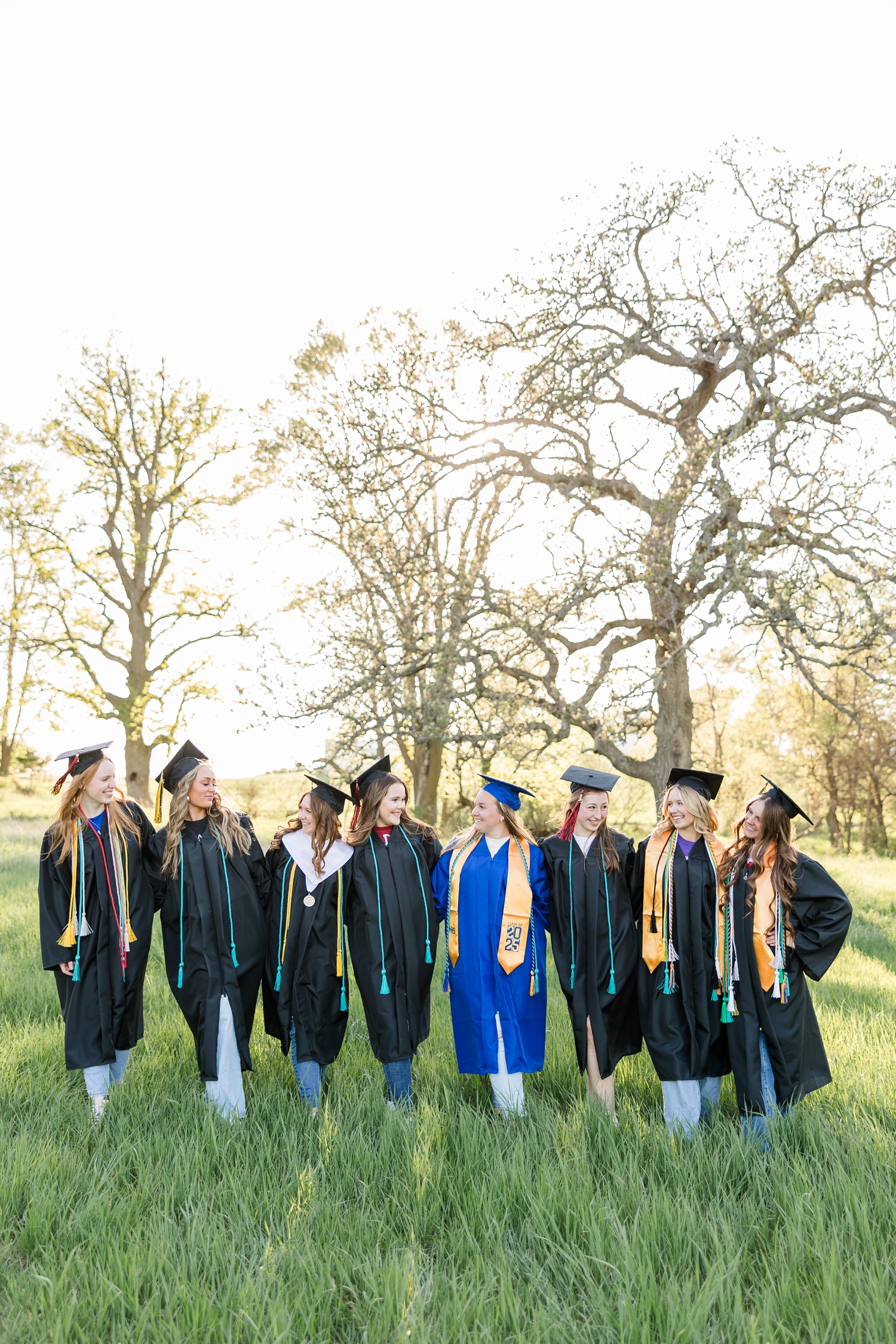 TEAM 25, senior photography spokesmodels, walk in a grass pasture during golden hour wearing their caps, gowns, stoles and cords, smiling and laughing at each other | CB Studio