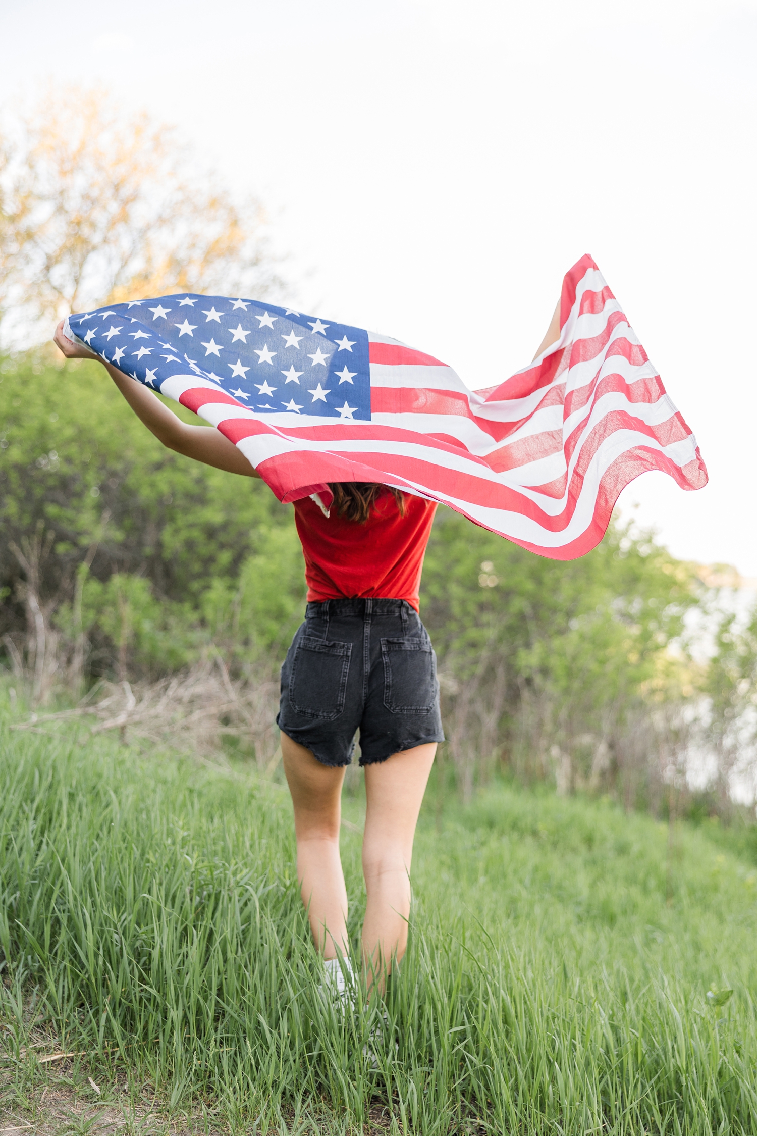 Senior, Danica, holds the American flag up behind her as it blows in the wind, while standing in a grass field | CB Studio