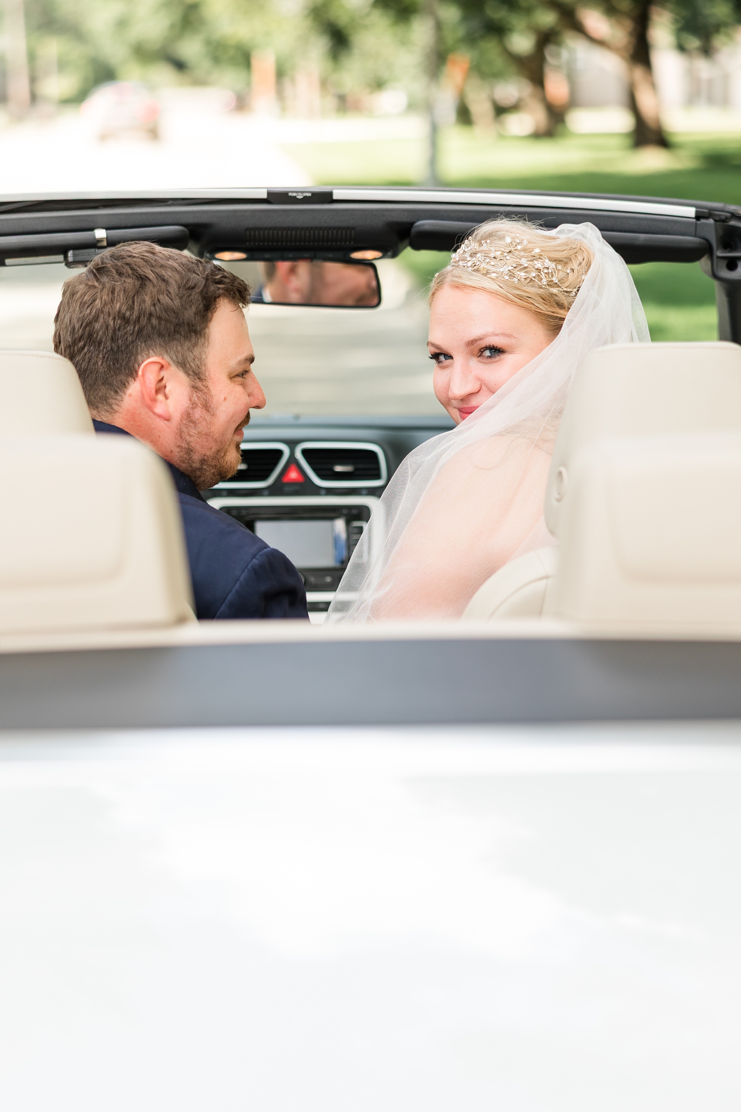 Natalie peeks over her shoulder has her and Connor sit in a white convertible, Connor smiles at Natalie | CB Studio