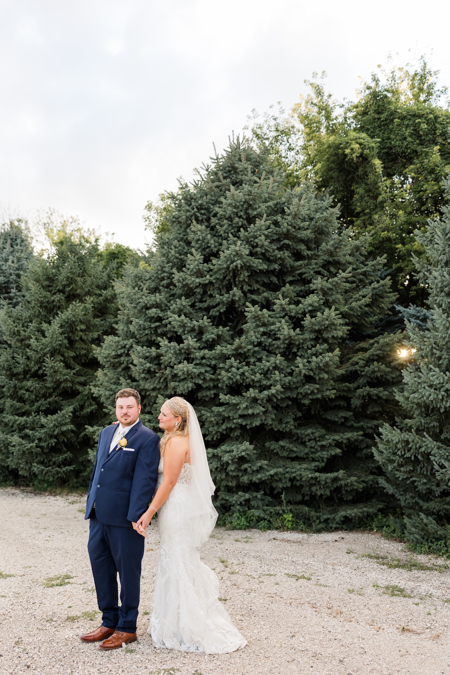 Natalie stands behind Connor and they hold hands as they stand in front of an evergreen tree line at sunset. Natalie smiles at Connor while he looks ahead stoically | CB Studio