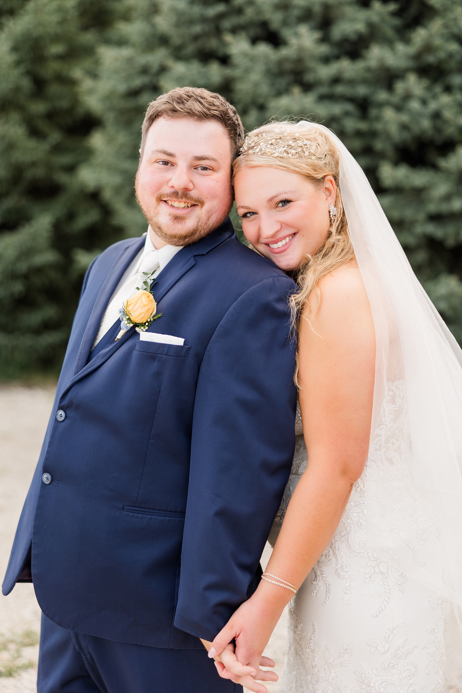 Natalie stands behind Connor and they hold hands as they stand in front of an evergreen tree line at sunset. Natalie rests her chin on Connor's shoulder as they both smile | CB Studio