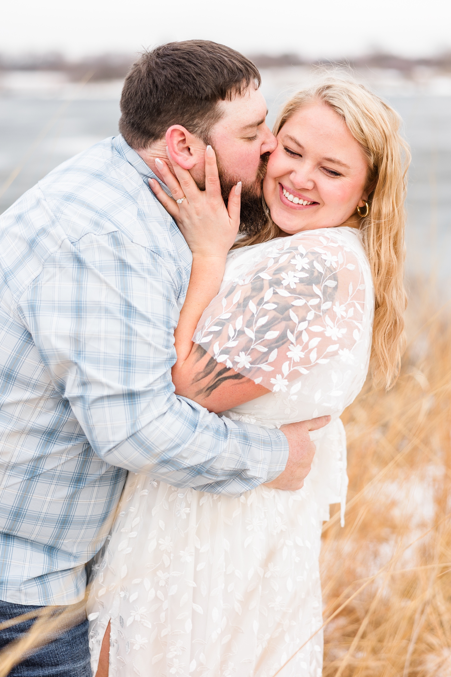 Teri grasps Dalton's face has he pulls her in and kisses her check with Smith Lake in the background at Water's Edge Nature Center during early spring in Iowa | CB Studio