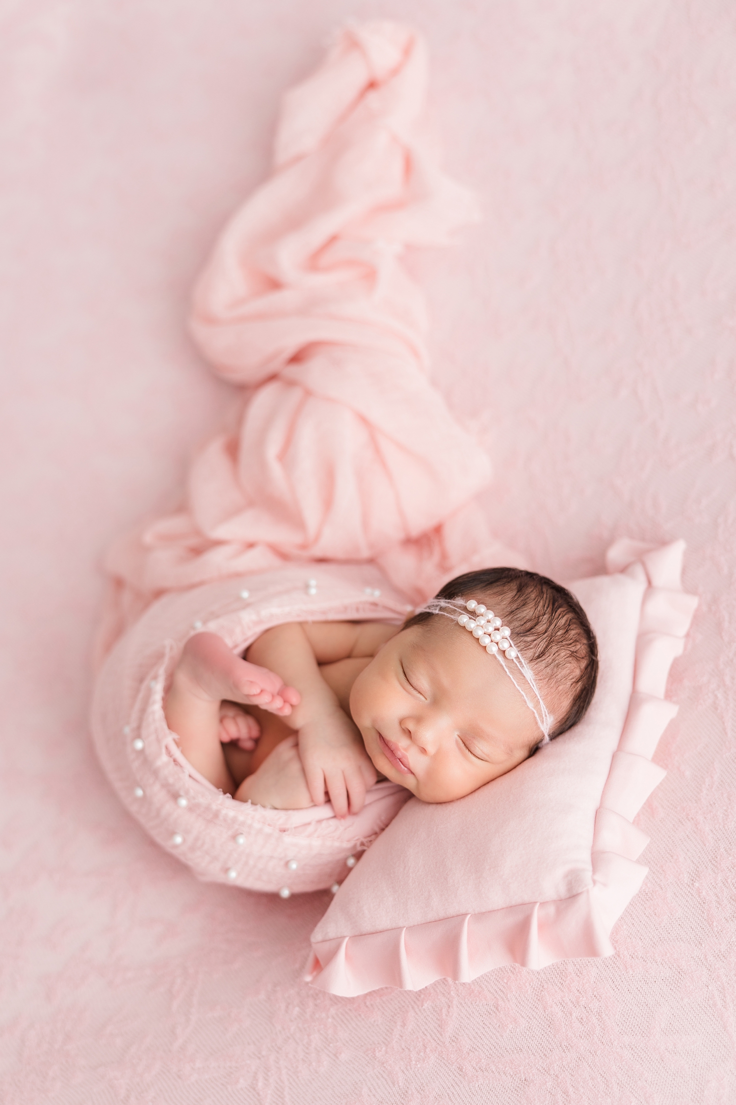 Baby Zayla adorned with a pearl headband and wrapped in pink with pearls on the top portion of the wrap and nestled on a floral lace pink backdrop and a pink ruffle pillow | CB Studio