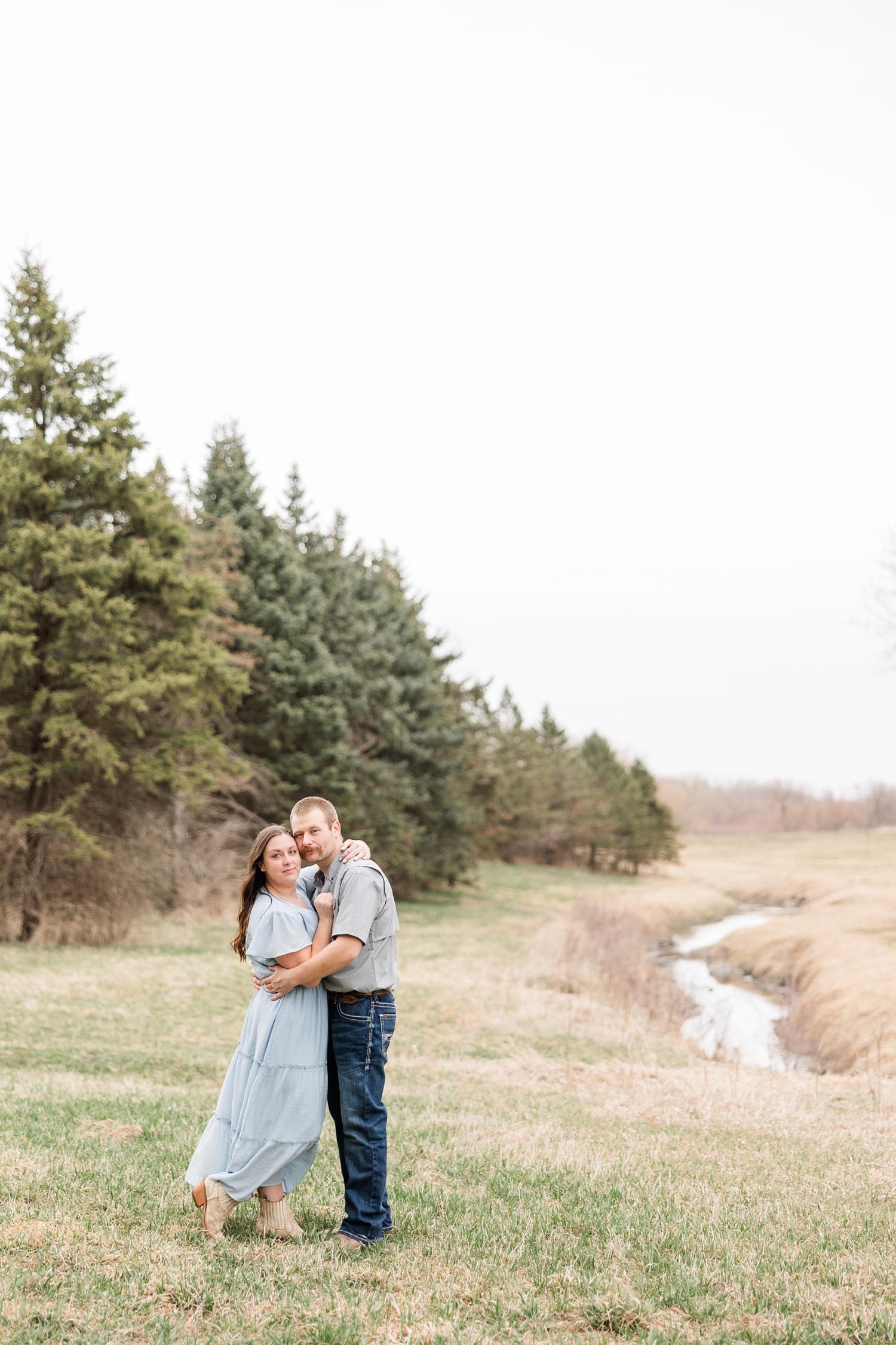 Ryan and Cassie embrace and lean their heads together with a flowing stream and a line of evergreen trees in the background | CB Studio