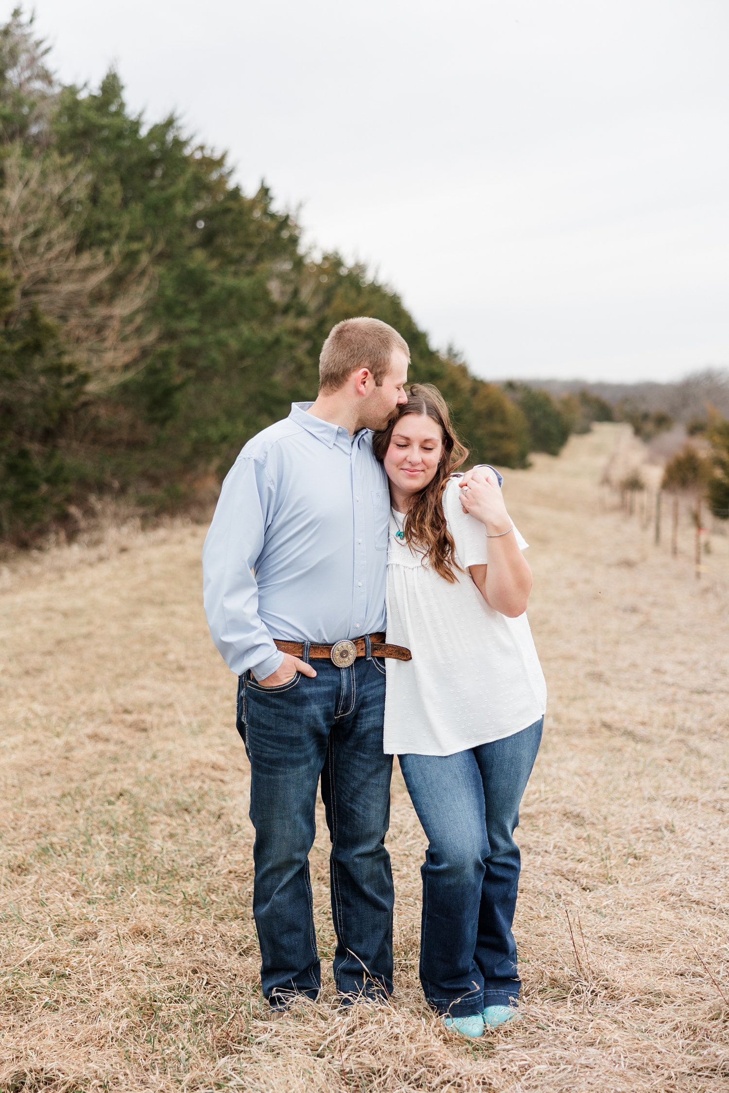 Ryan pulls Cassie in close by her shoulder and kisses the top of her head with rolling hills, a fence line, and evergreen trees in the background | CB Studio