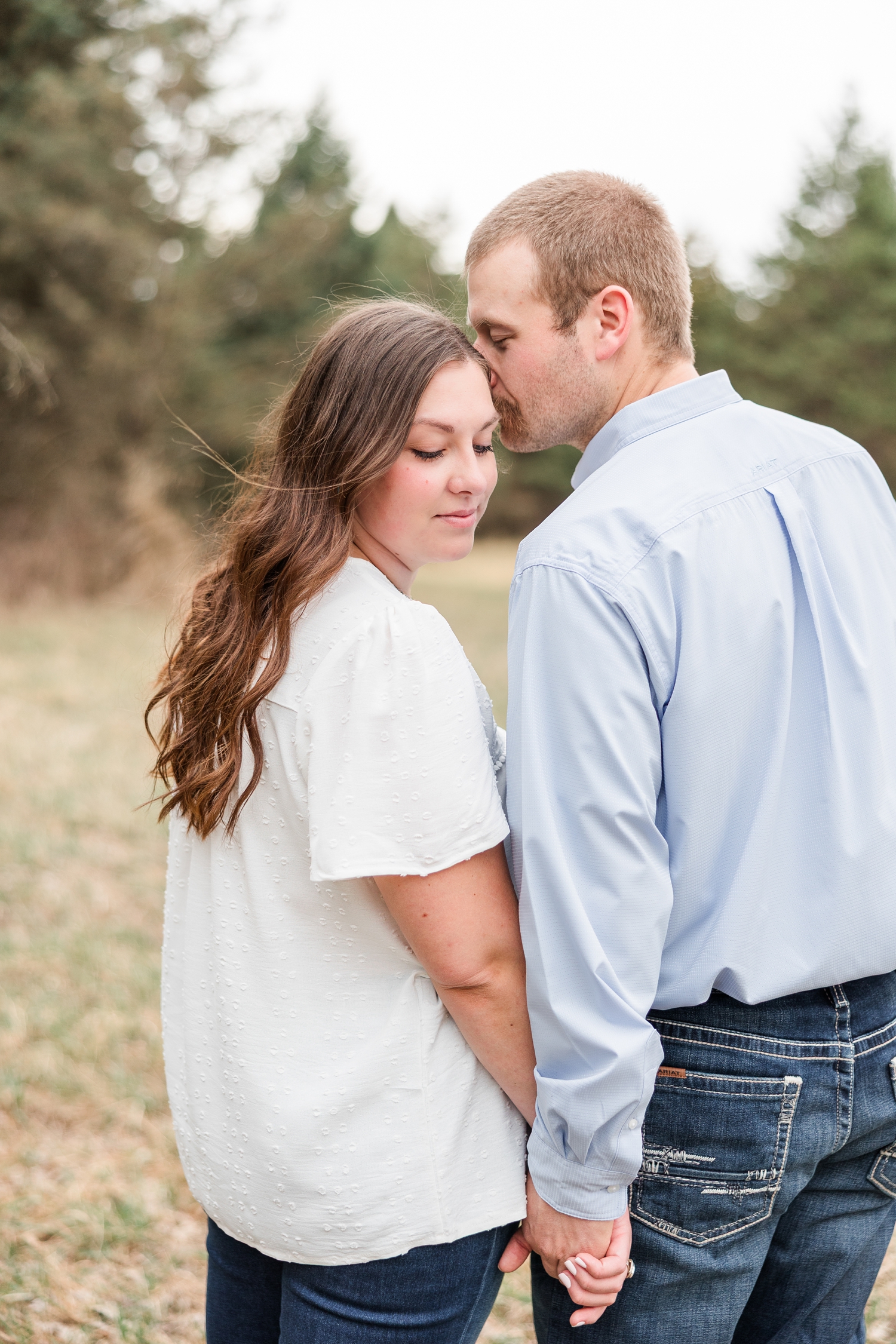 Ryan holds Cassie's hand and nuzzles her temple as she looks down over her shoulder with evergreen trees surrounding the background | CB Studio
