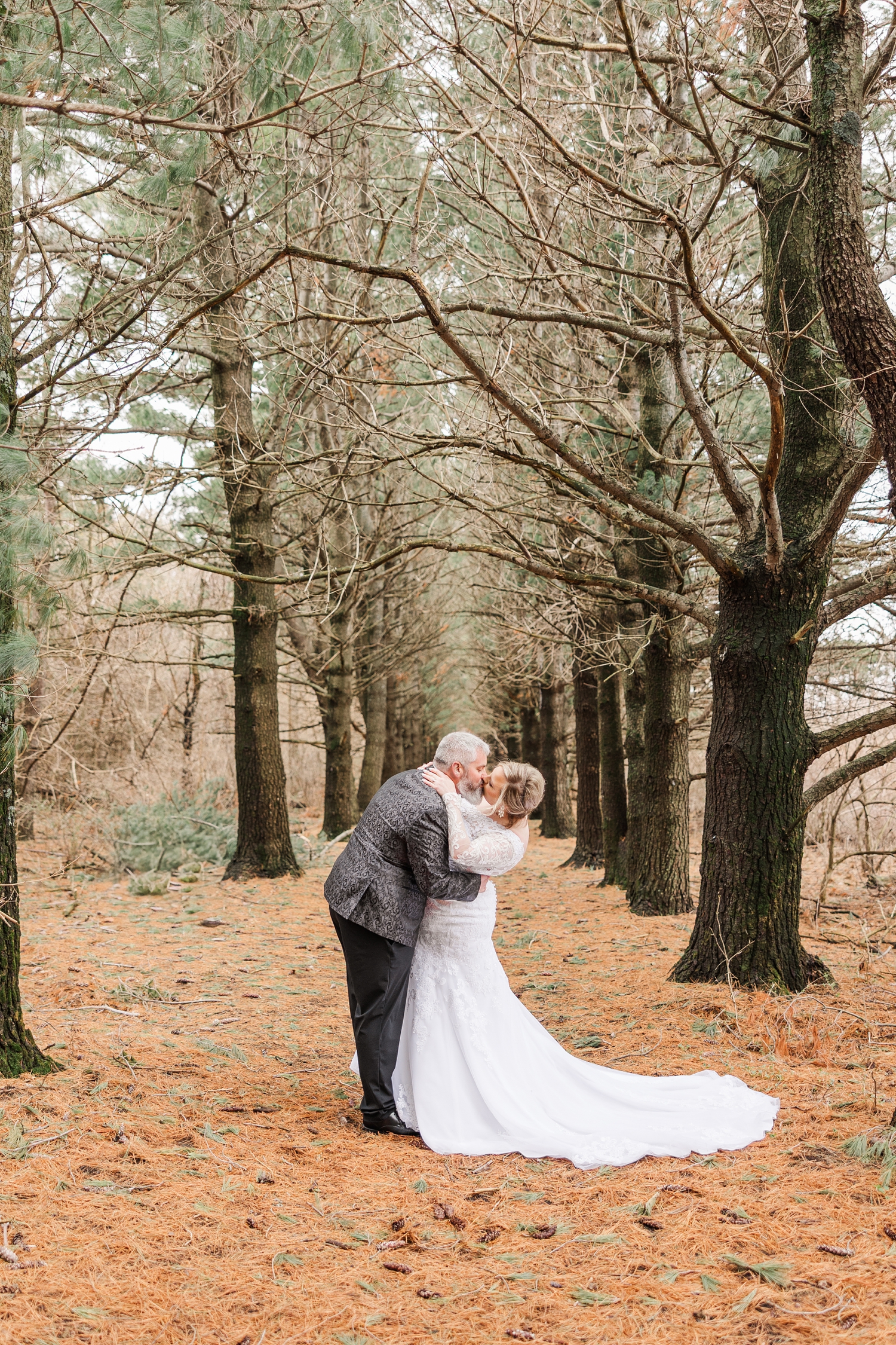 Nick dips Sydney back and kisses her as they stand in the middle of the narrow pathway beneath the pines of the Water's Edge Nature Center | CB Studio