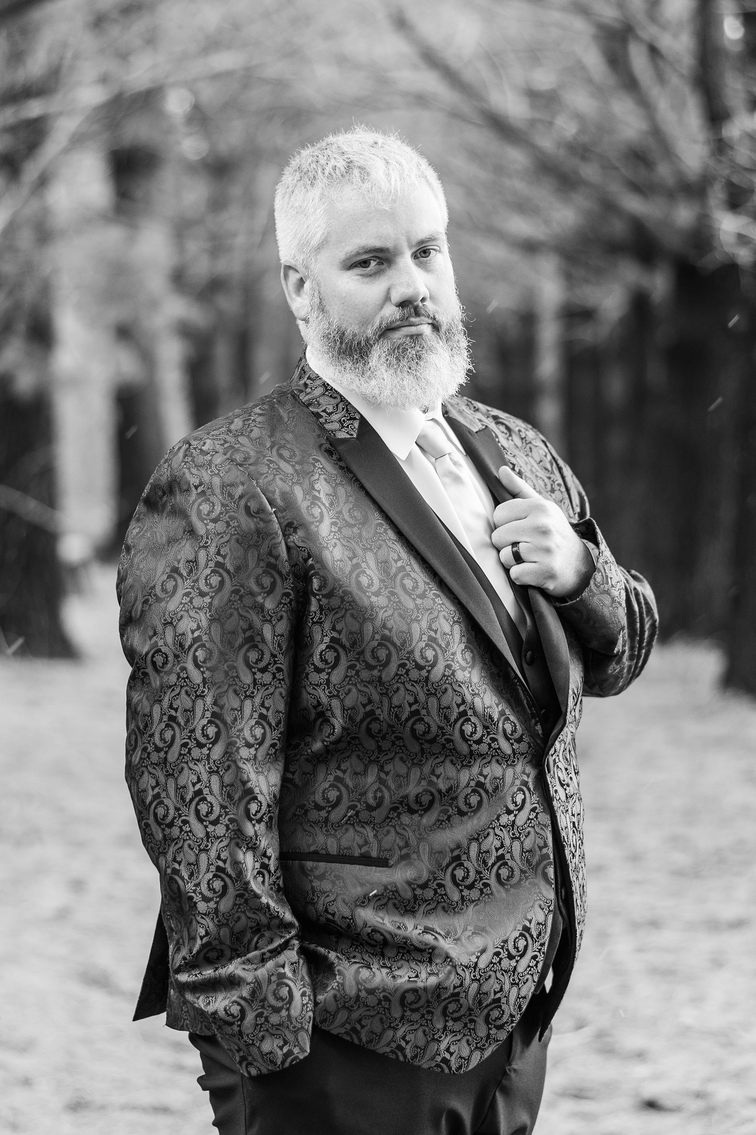 Nick stands stoically with one hand in his pocket and the other holding his lapel in the middle of the narrow pathway beneath the pines of the Water's Edge Nature Center | CB Studio