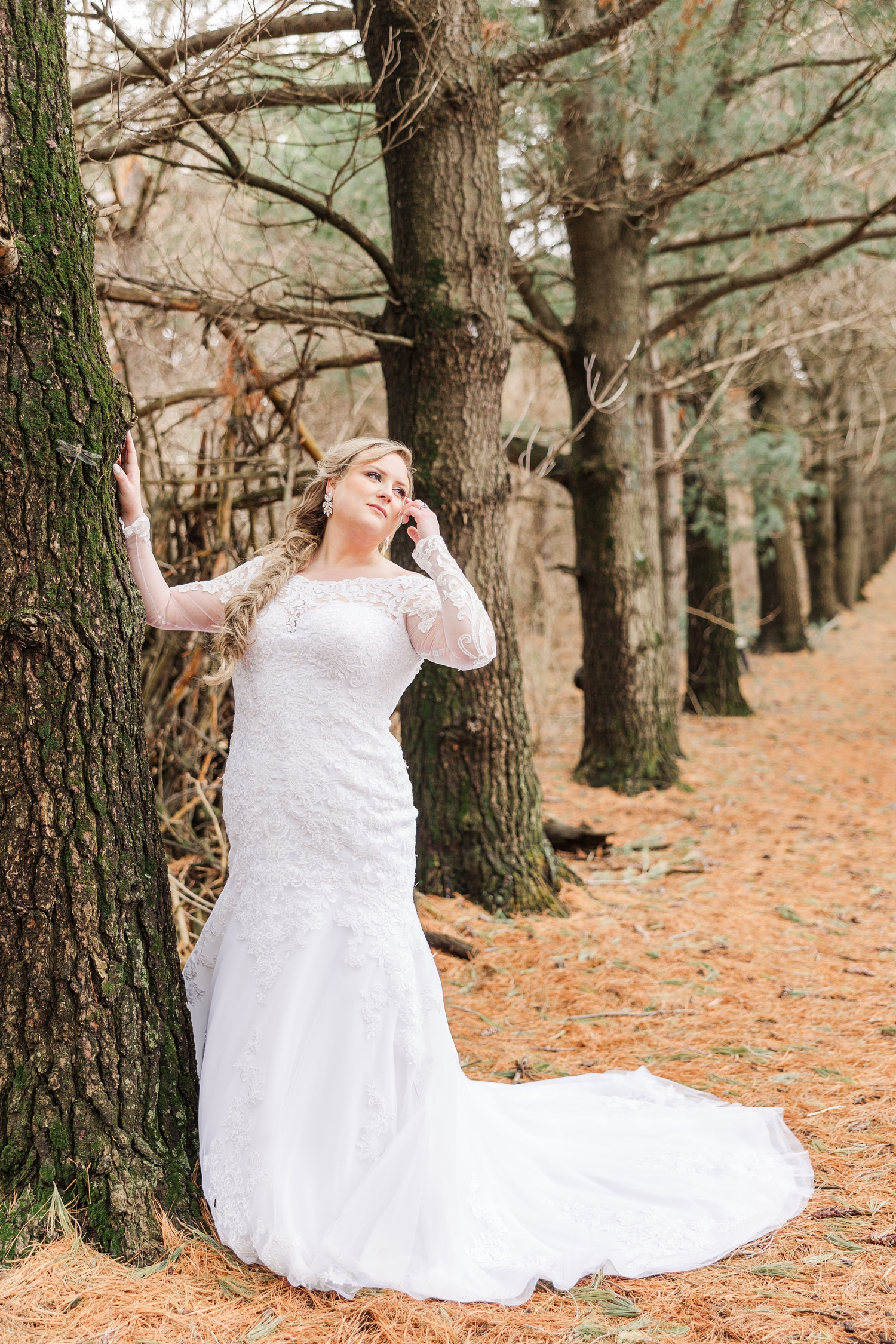 Sydney leans against a pine tree as she looks up and tuck her hair behind her ear in the middle of the narrow pathway beneath the pines of the Water's Edge Nature Center | CB Studio