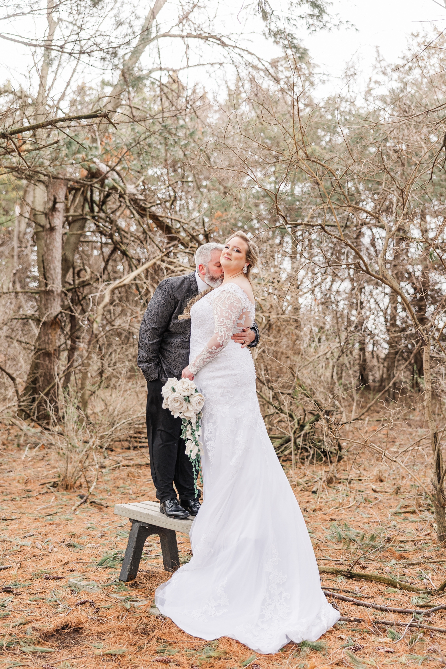 Nick slowly kisses Sydney's neck as they stand on top of a bench beneath the pines of the Water's Edge Nature Center | CB Studio