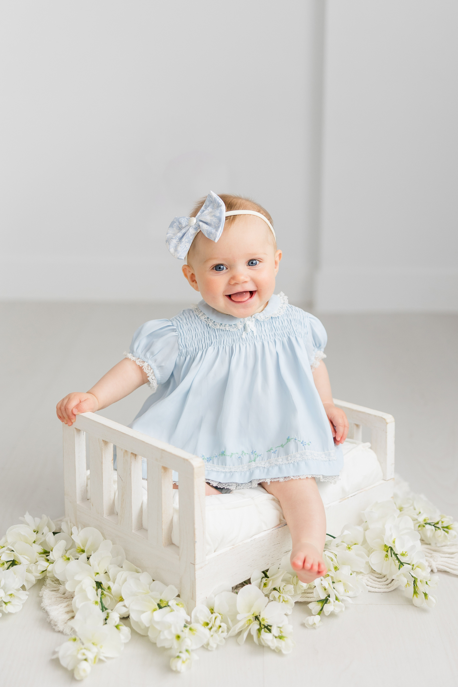 Baby Adalyn is all smiles as she sits in a small white doll bed surrounded with white flowers in an all white room as she wears a light blue vintage baby dress that once belonged to her grandmother | CB Studio