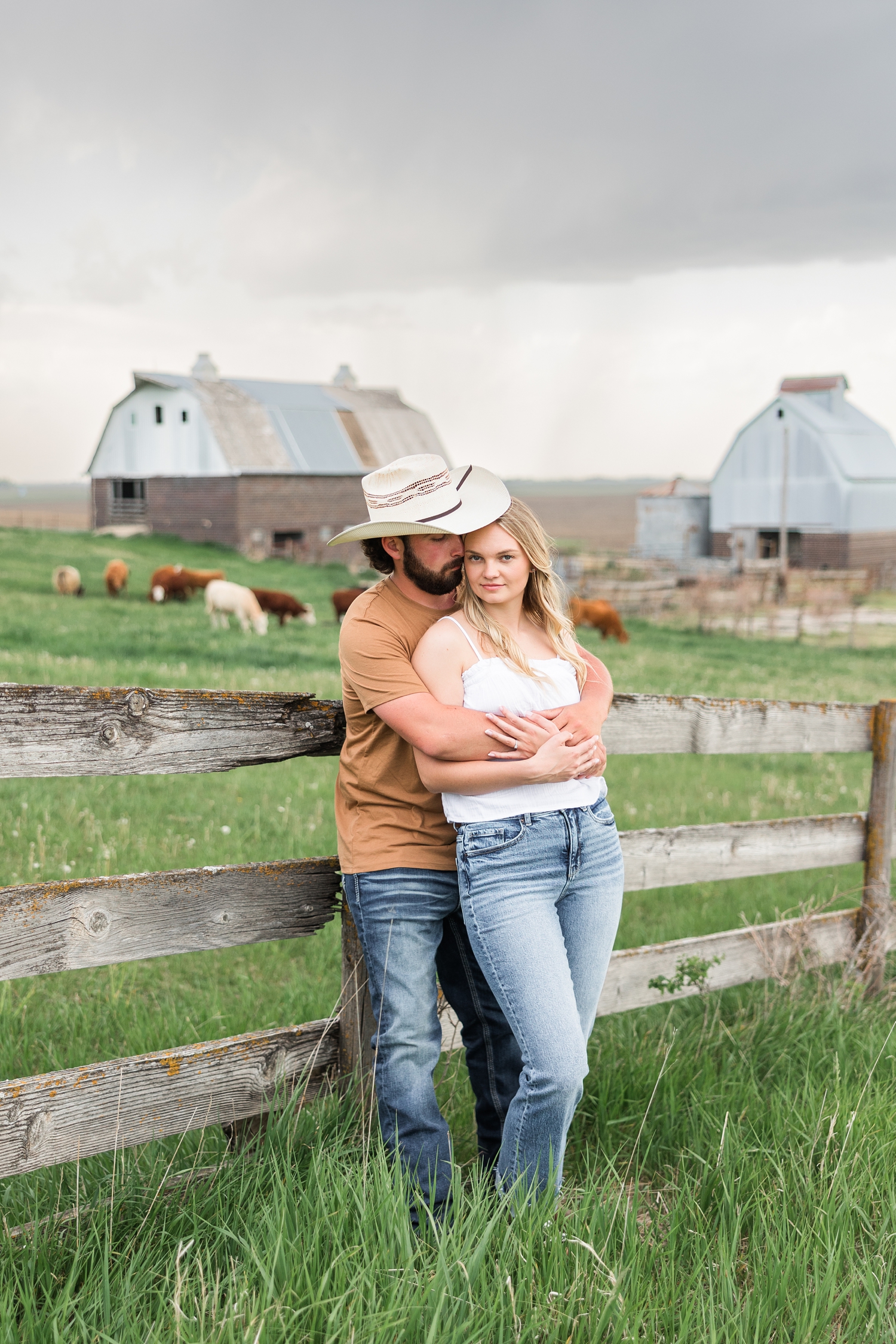 Dawson embraces Jade from behind and nuzzles her as they both lean against an old wooden fence in the middle of an Iowa cattle pastures with cows and storm clouds in the background | CB Studio