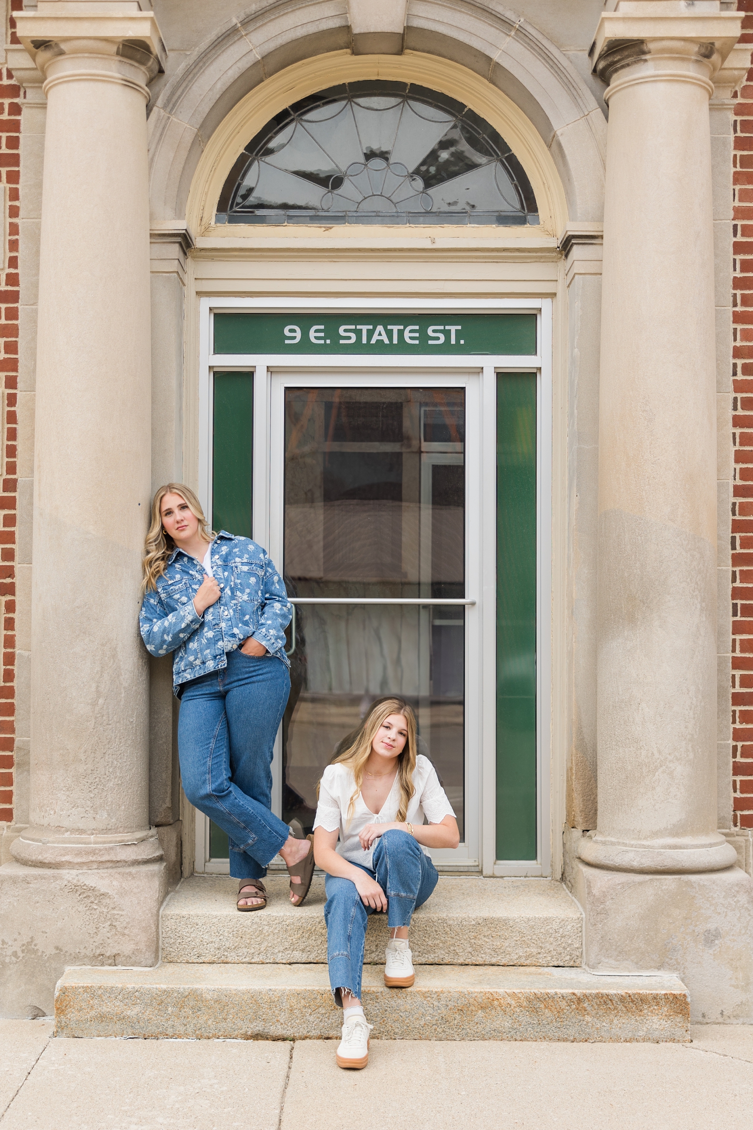 Ocean and Mya dressed in denim styles, pose in front of a old stone building with tan columns framing the door in Algona, IA | CB Studio