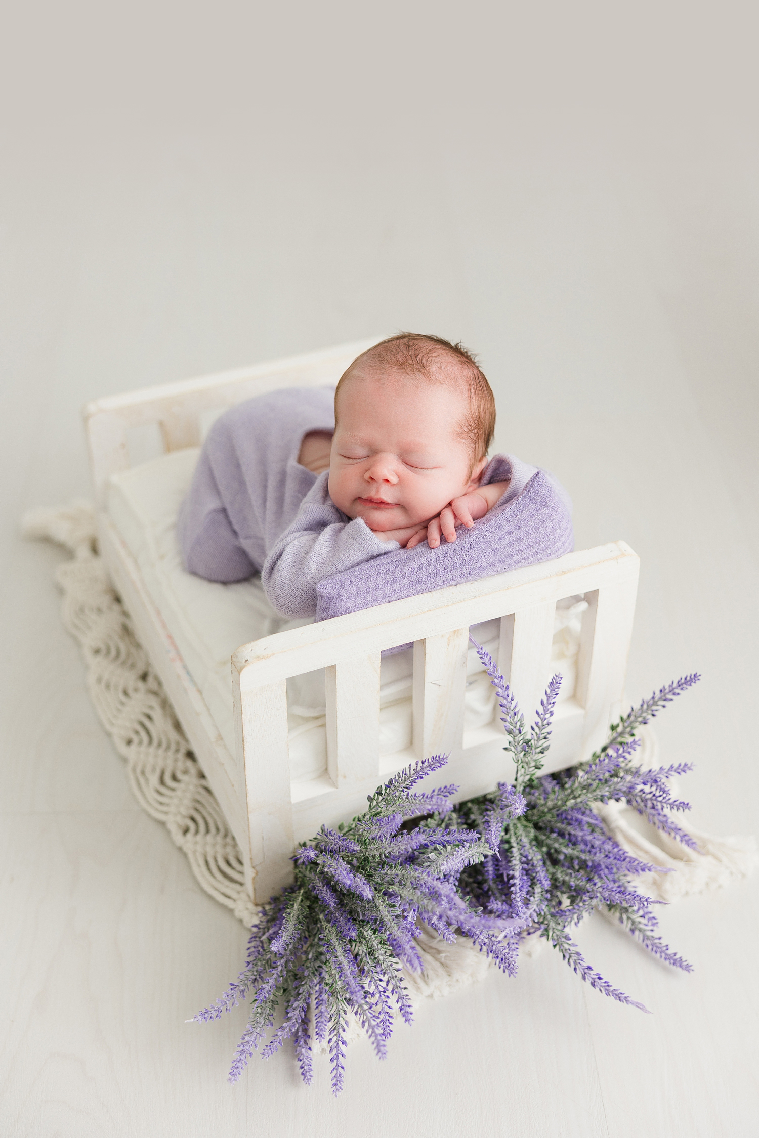 Baby Ella, in a soft, lavender newborn sleeper, is posed with her chin resting on her arms that are resting on top of a lavender pillow nestled in a white wooden doll bed surrounded with lavender florals on a white wood floor | CB Studio