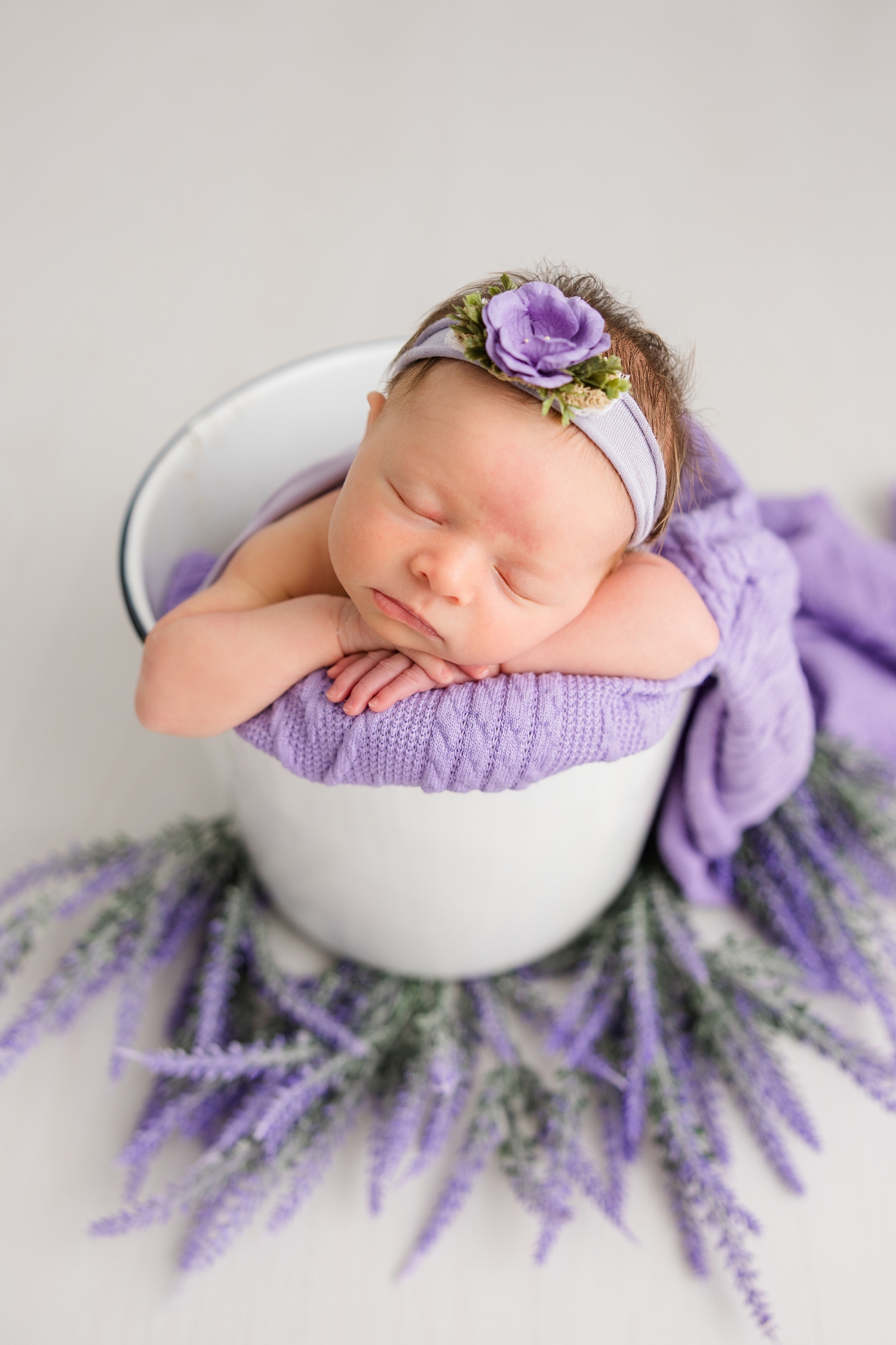 Baby Sophia, wearing a purple floral headband, is nestled in a white bucket with purple fabric on top of a white floor with the bucket surrounded with lavender sprigs | CB Studio