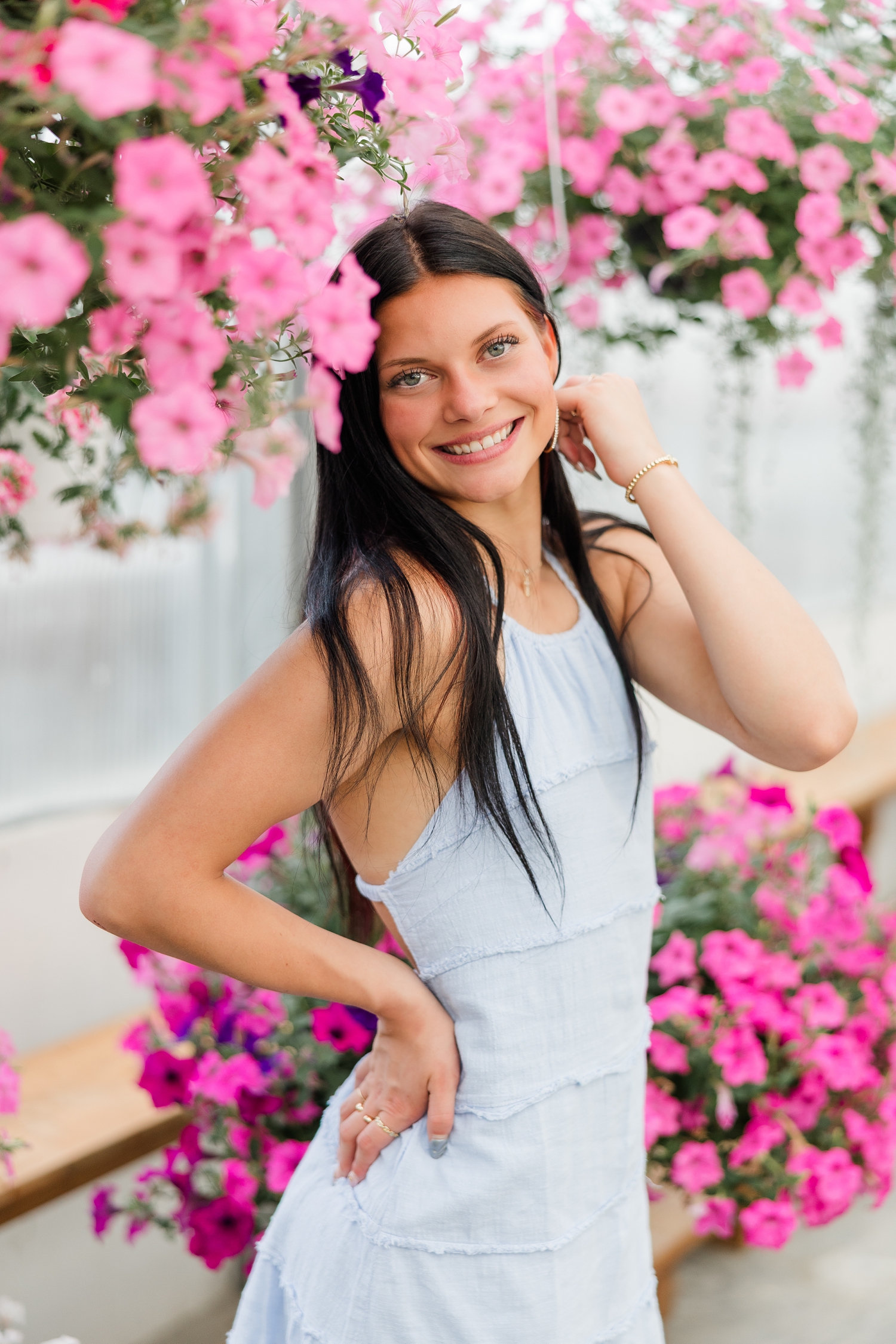 Brooke, wearing a light blue linen dress, smiles as she is surrounded by pink petunias at Cottage Gardens while she has one hand on her hip and the other tucks hair behind her ear | CB Studio