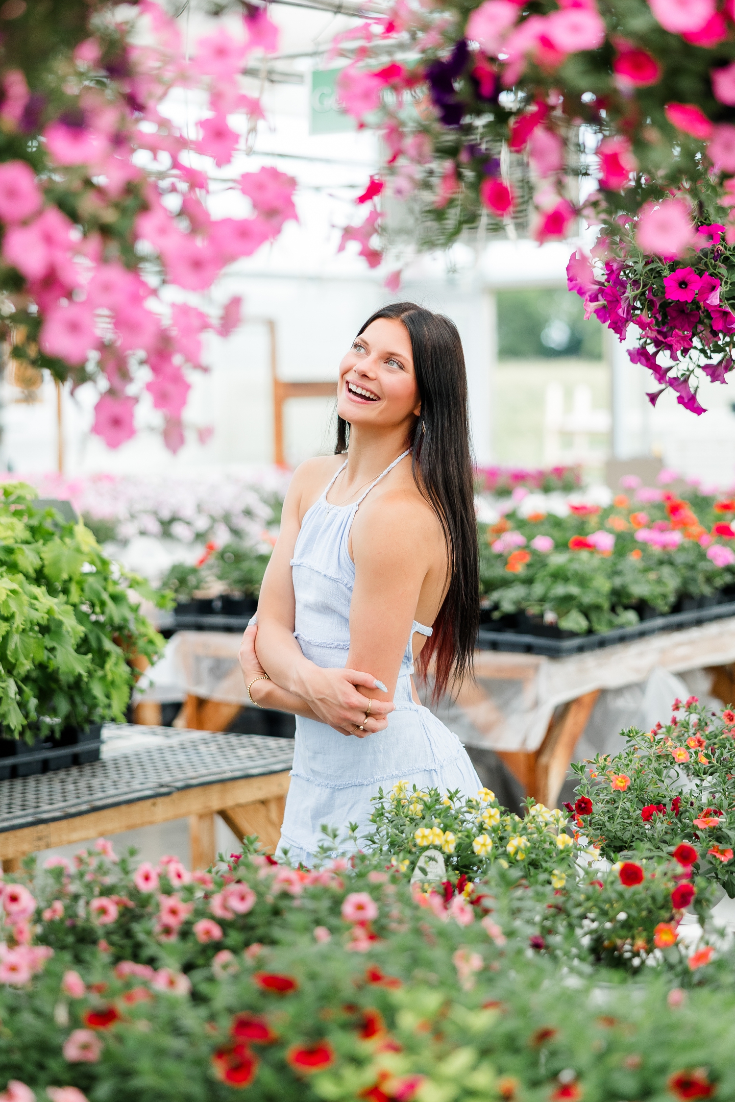 Brooke, wearing a light blue linen dress, smiles as she is surrounded by pink and multi-colored petunias at Cottage Gardens while she crosses her arms and laughs while looking up | CB Studio