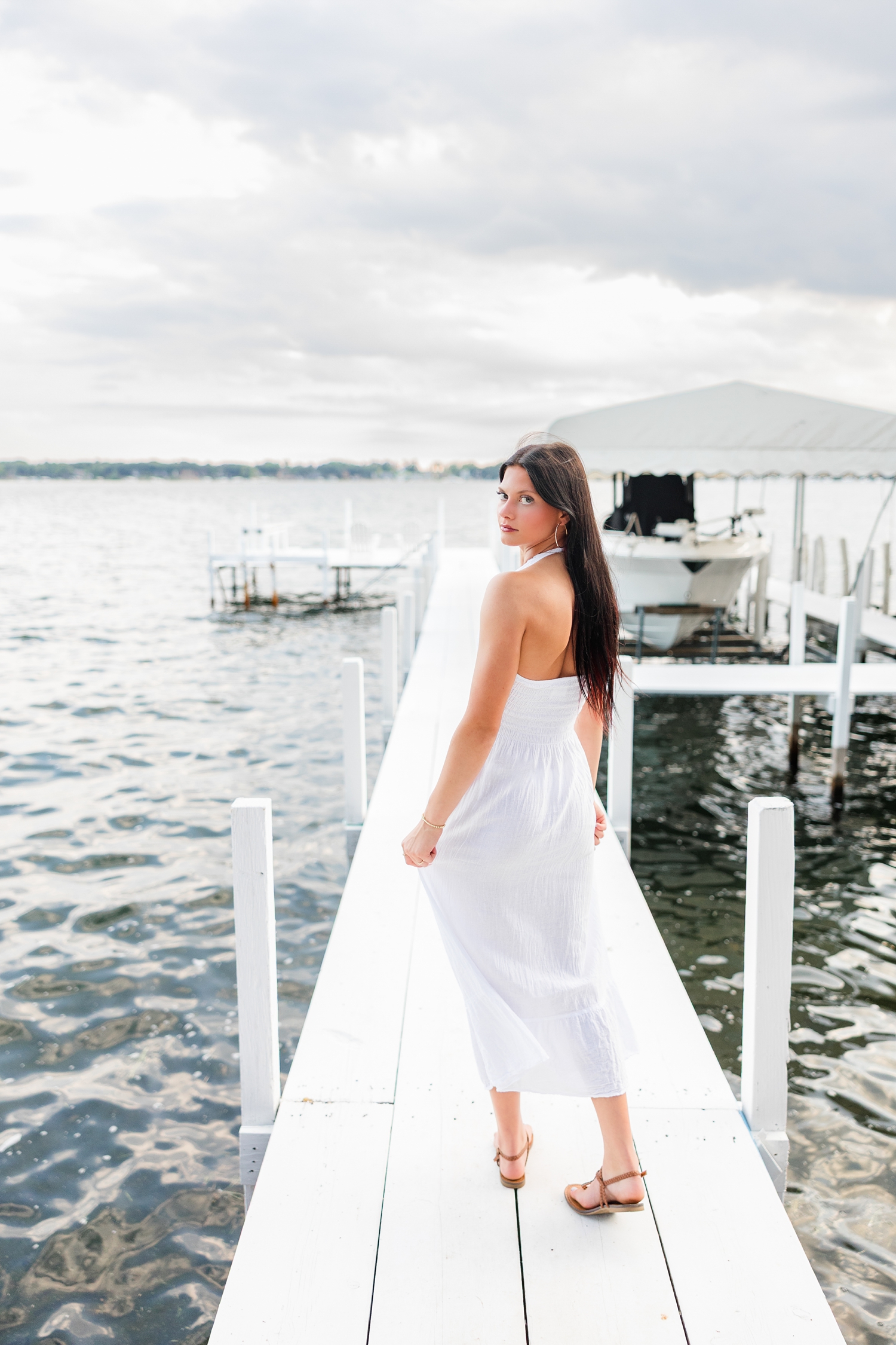 Brooke, wearing a long white linen dress, looks back as she is walking down an all white dock on West Okoboji Lake | CB Studio