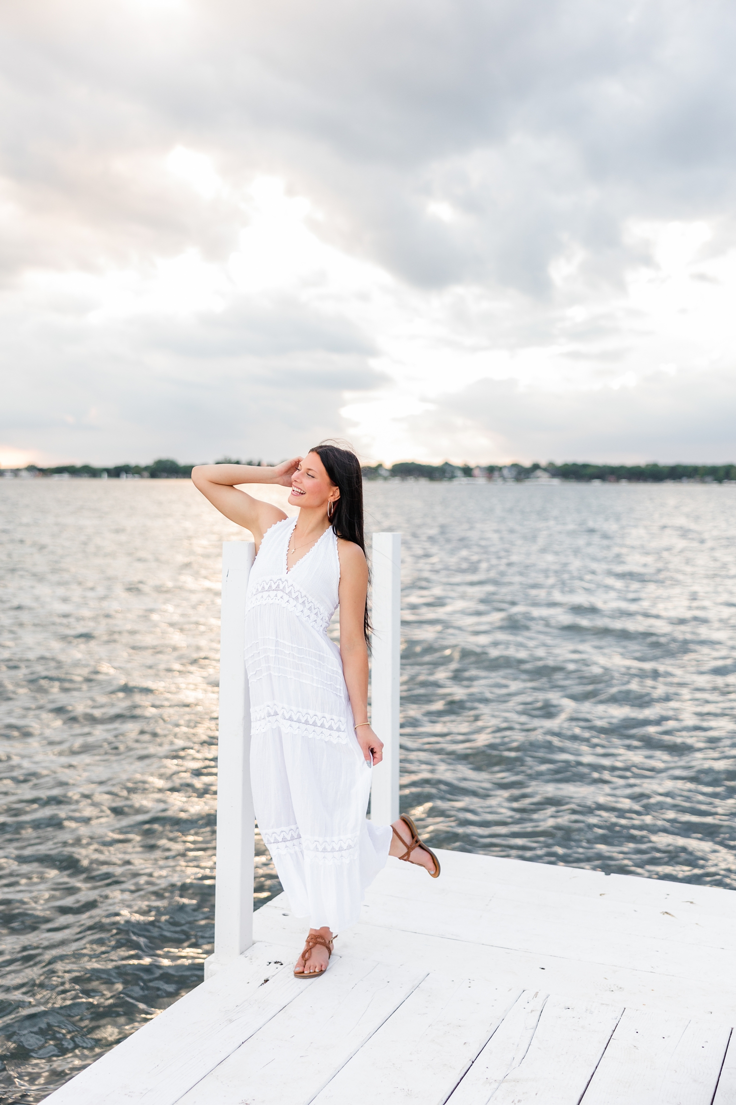 Brooke, wearing a long white linen dress, leans on a post of an all white dock on West Okoboji Lake | CB Studio
