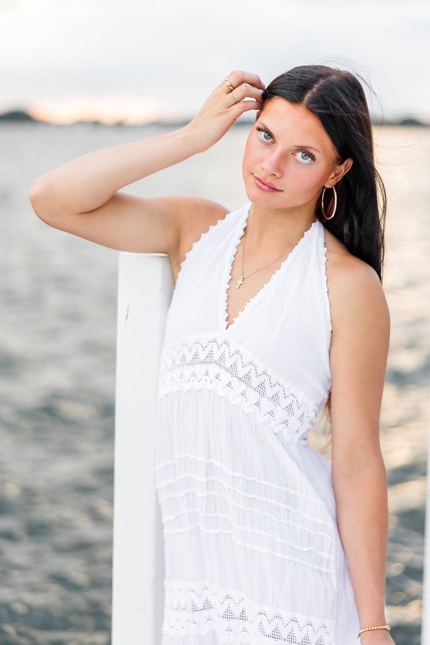 Brooke, wearing a long white linen dress, leans on a post of an all white dock on West Okoboji Lake as she tucks her hair behind her ear | CB Studio