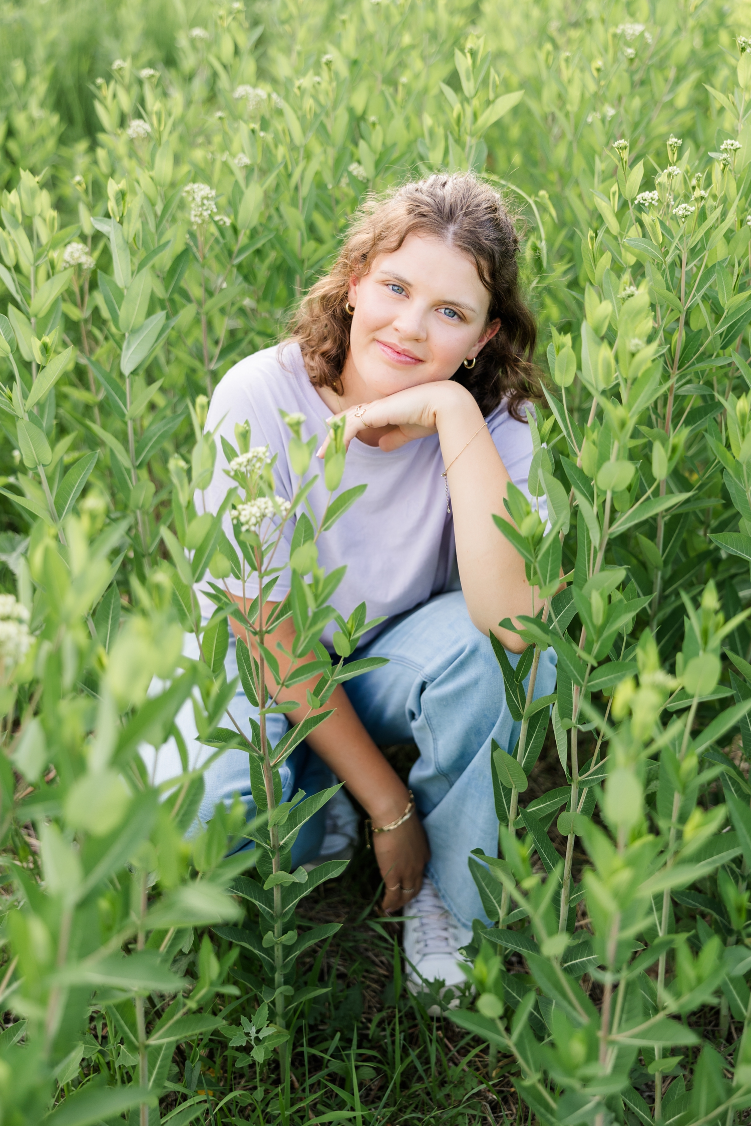 Ellie, wearing a purple t-shirt and jeans, squats down in a grass pasture surrounded by green foliage | CB Studio