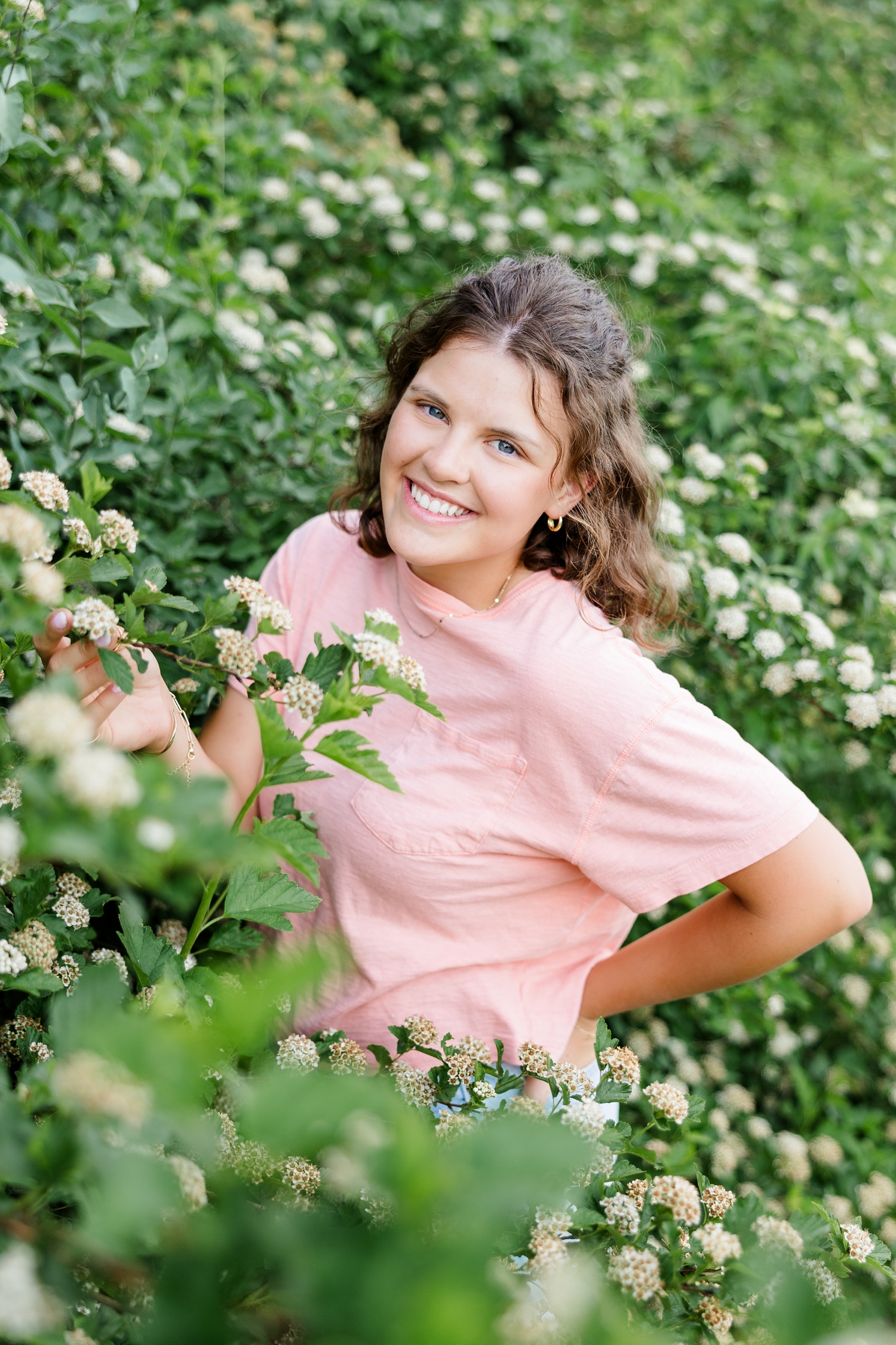 Ellie, wearing a t-shirt, stands in a patch of white floral bushes with her hand in her back pocket | CB Studio