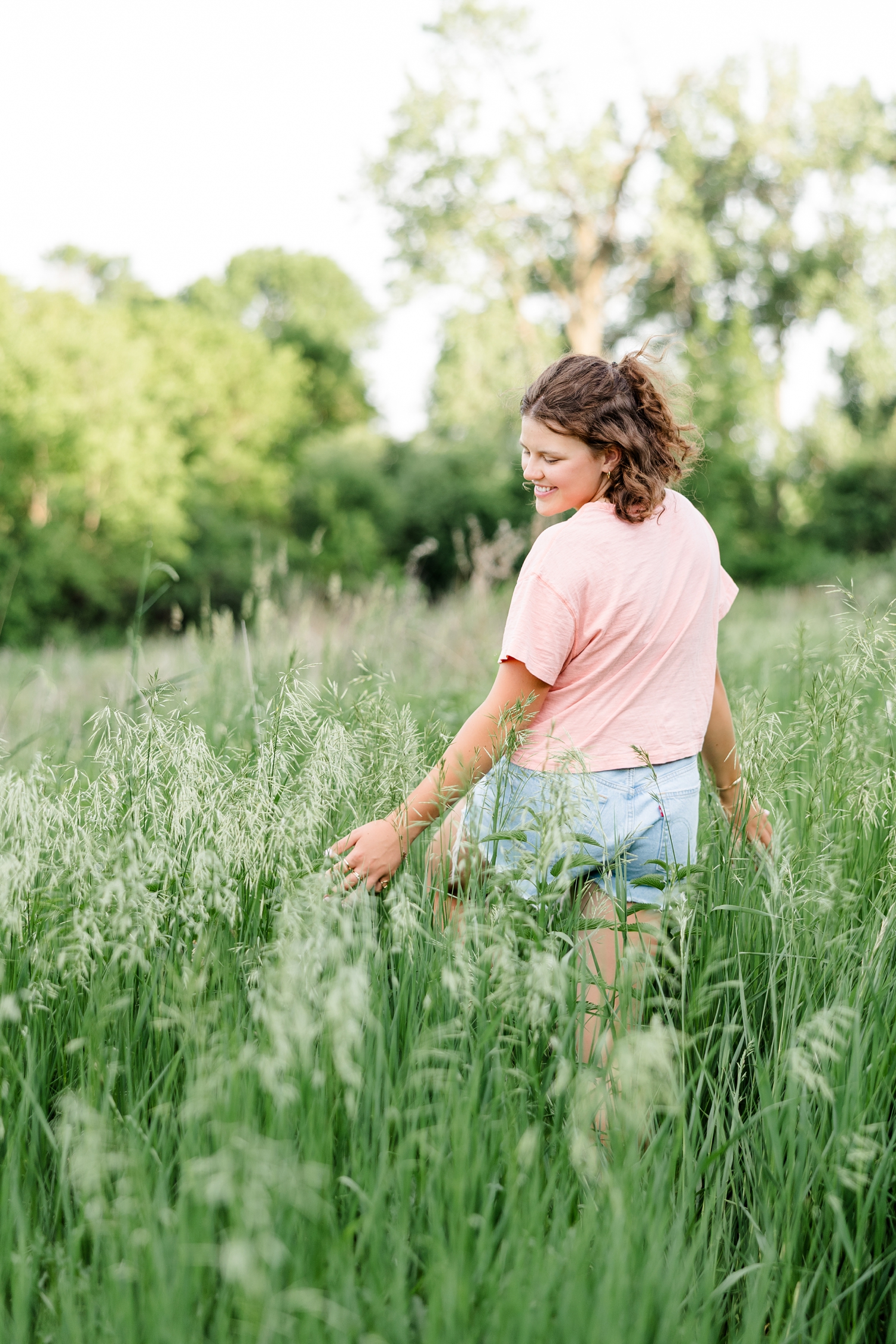 Ellie, wearing a t-shirt and denim shorts, walks away in a tall grass pasture as she brushes the grass with her hands and looks down while smiling | CB Studio