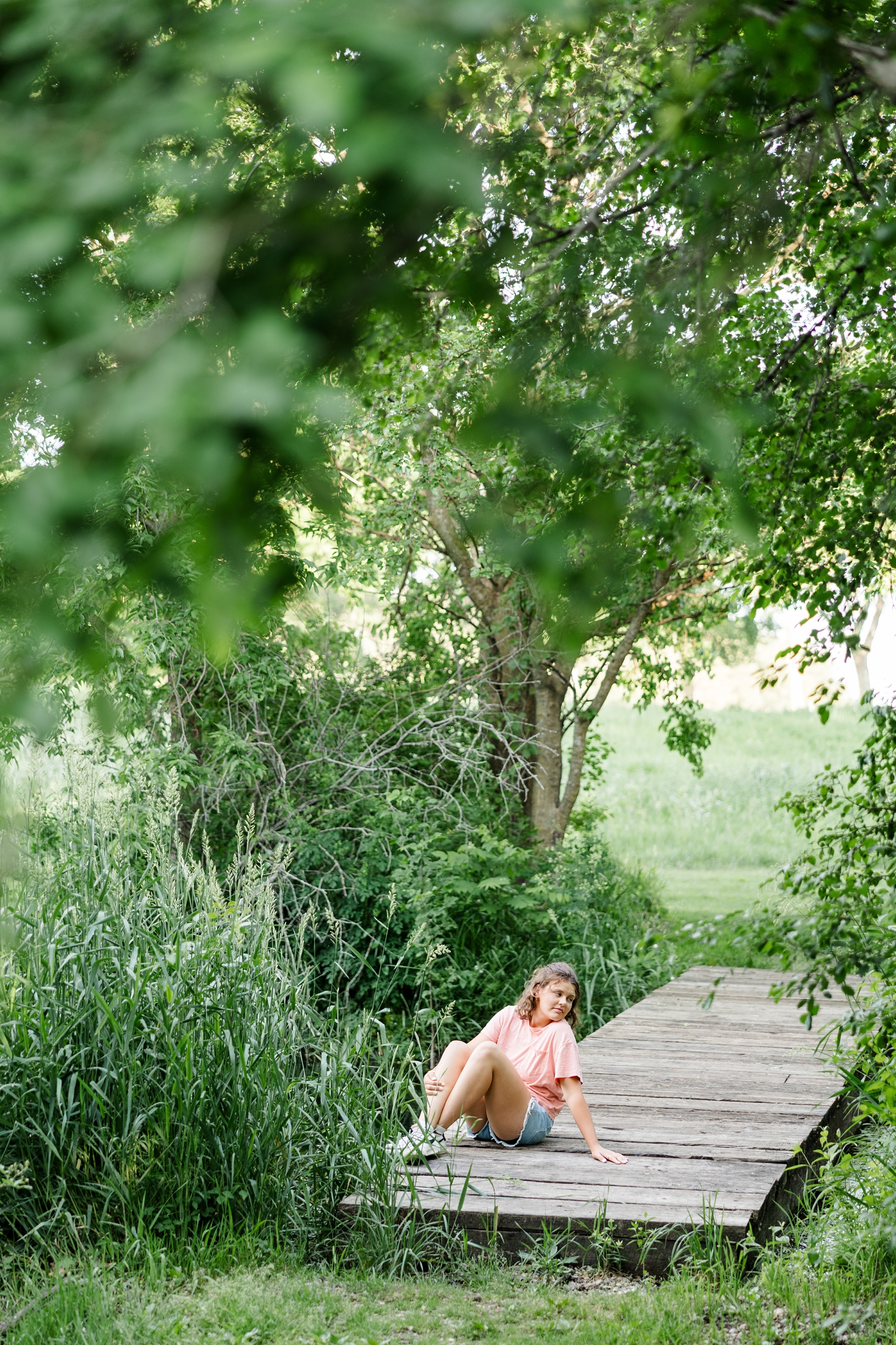 Ellie, wearing a t-shirt and denim shorts, sits on a wooden dock surrounded by trees with her knees up while she looks over her shoulder to the right | CB Studio