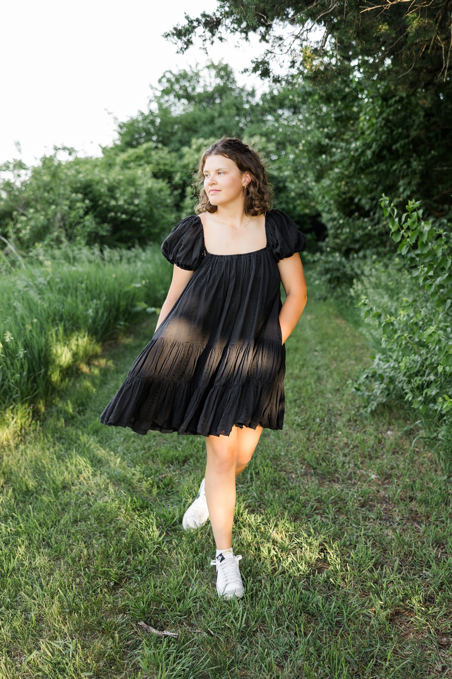 Ellie, wearing a black, flowing dress, walks along a grass path surrounded with trees and her dress flows and she looks to the left | CB Studio