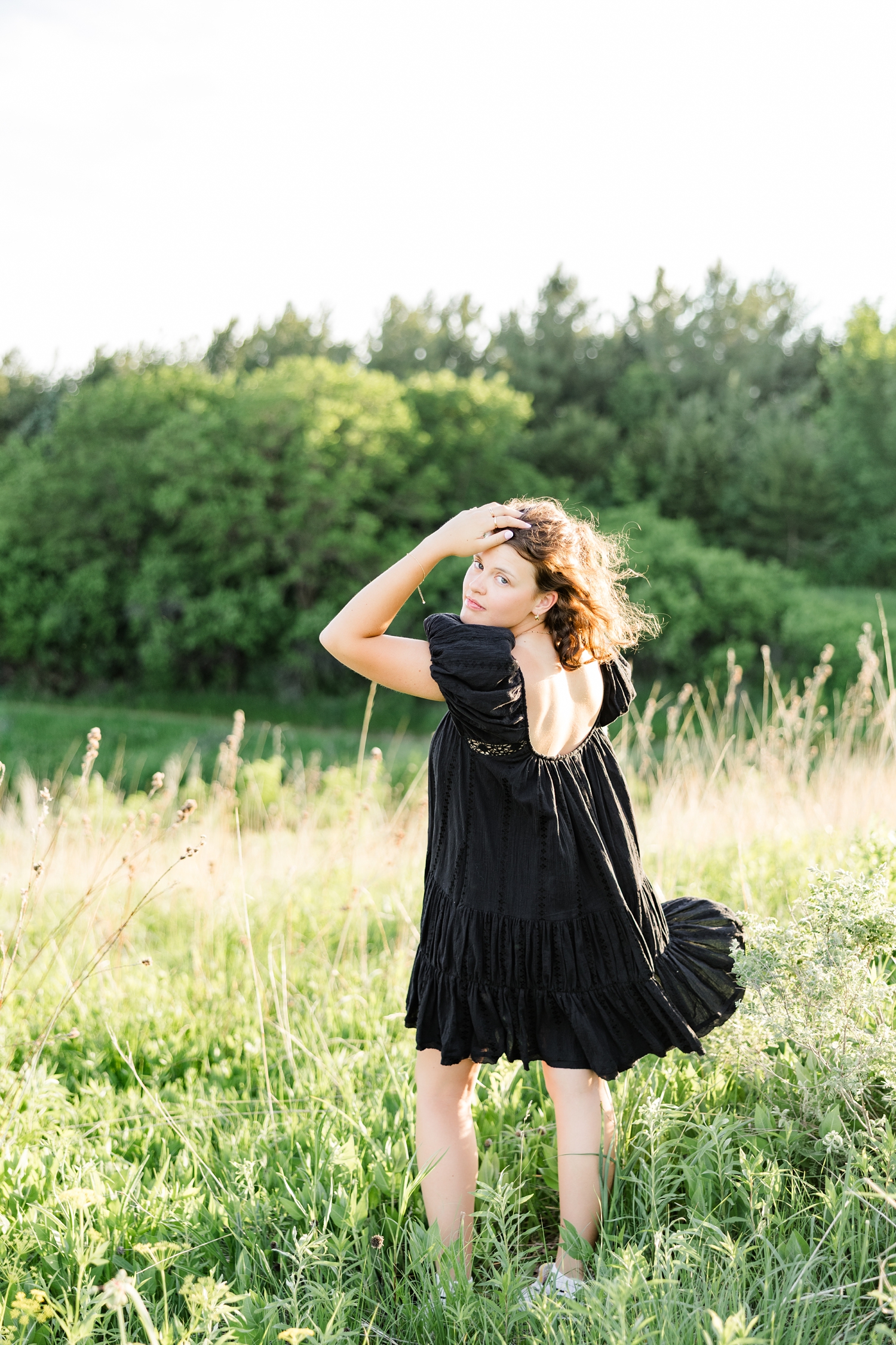 Ellie, wearing a black, flowing dress, stands in a grassy field as she looks over her shoulder back at the camera as the wind blows trough her dress and hair | CB Studio