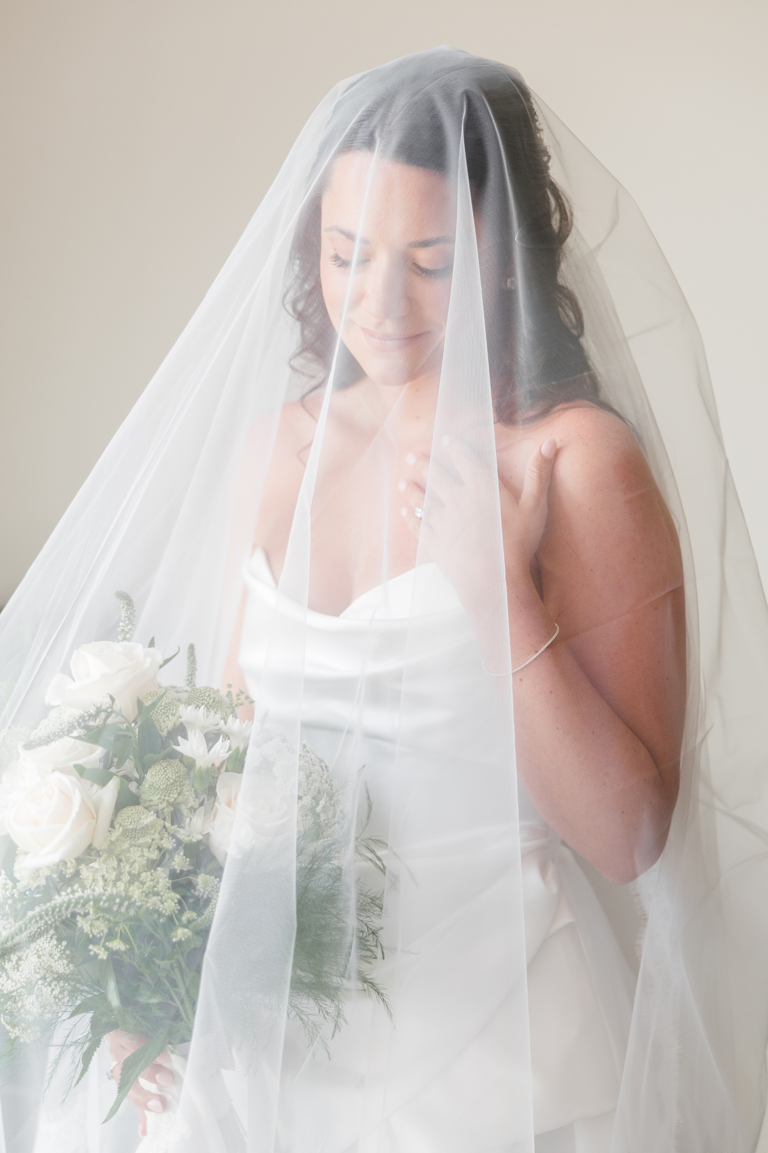 Bride, Emily, stands with her veil draped over her head and looks down at her wedding bouquet | CB Studio