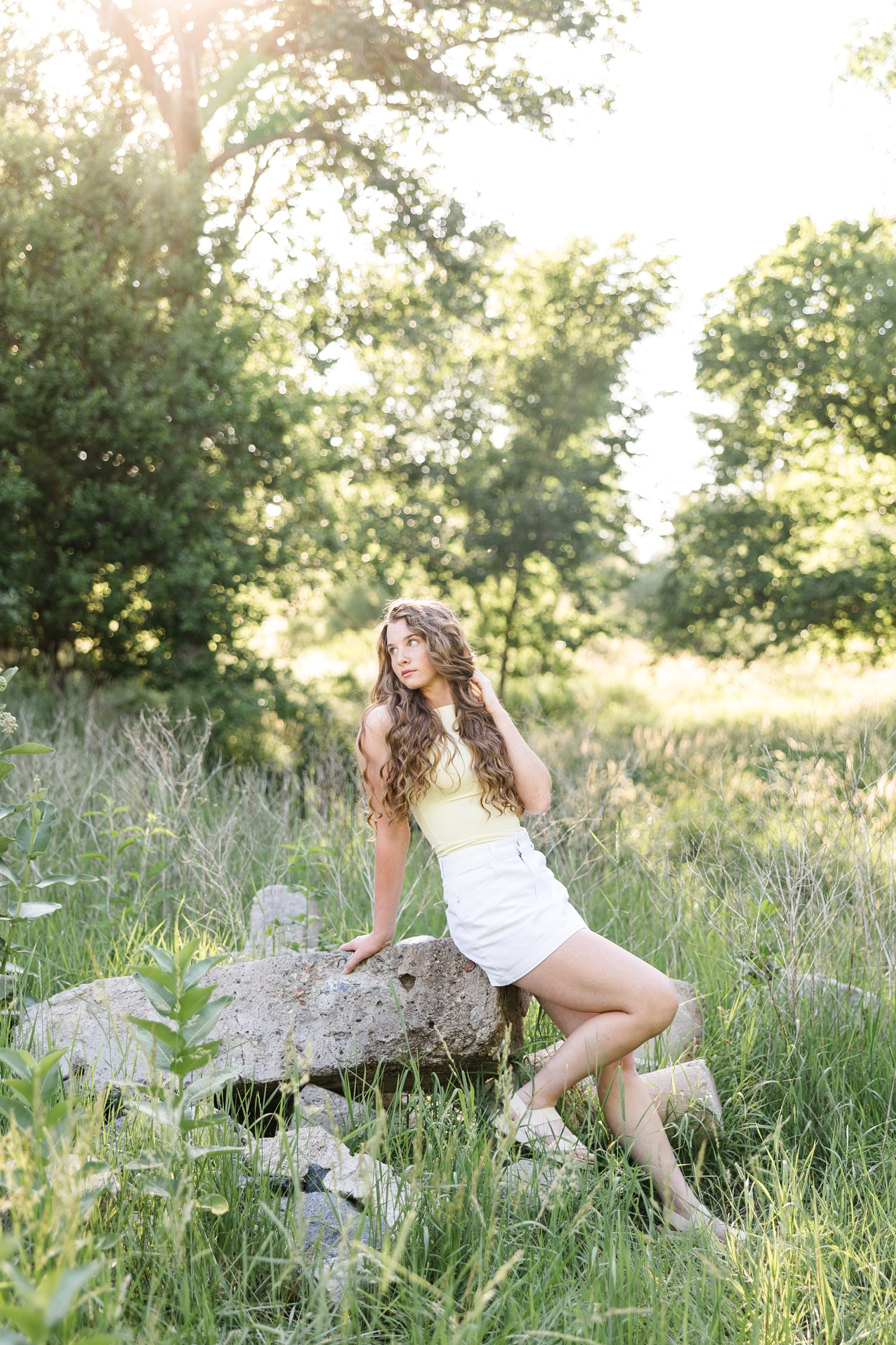 Mya, wearing a fitted yellow top and white denim skirt, sits on a rock, leans back and looks over her shoulder | CB Studio