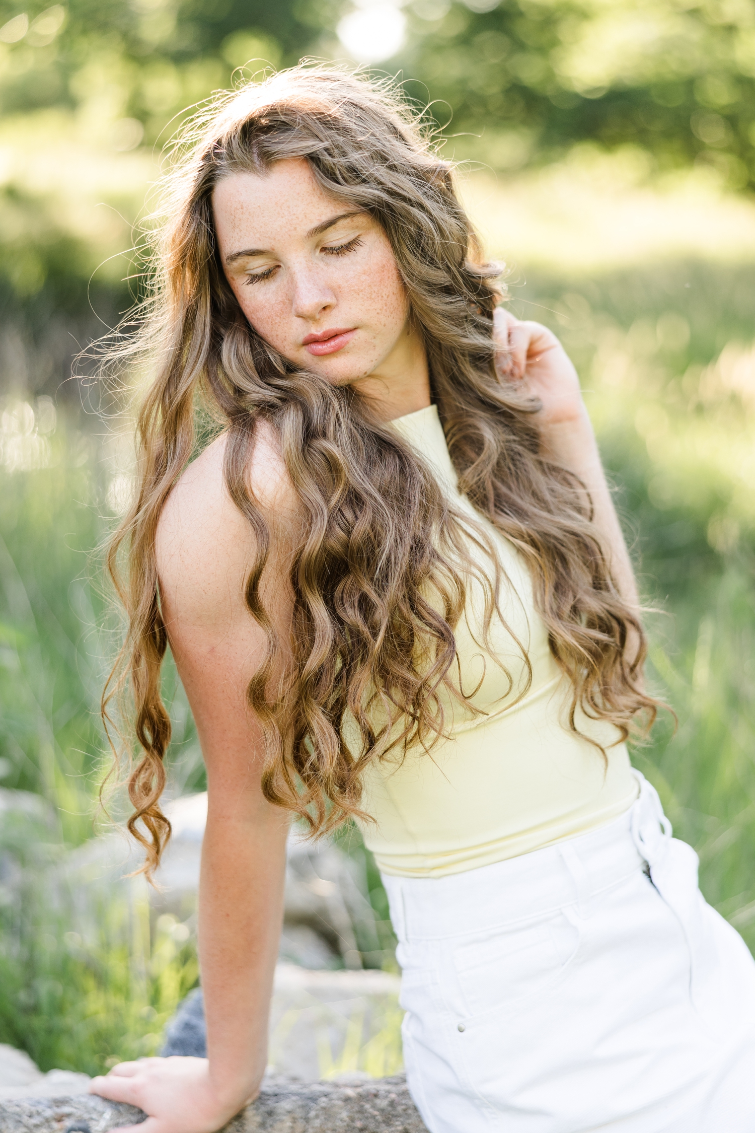 Mya, wearing a fitted yellow top and white denim skirt, sits on a rock, leans back and looks down | CB Studio