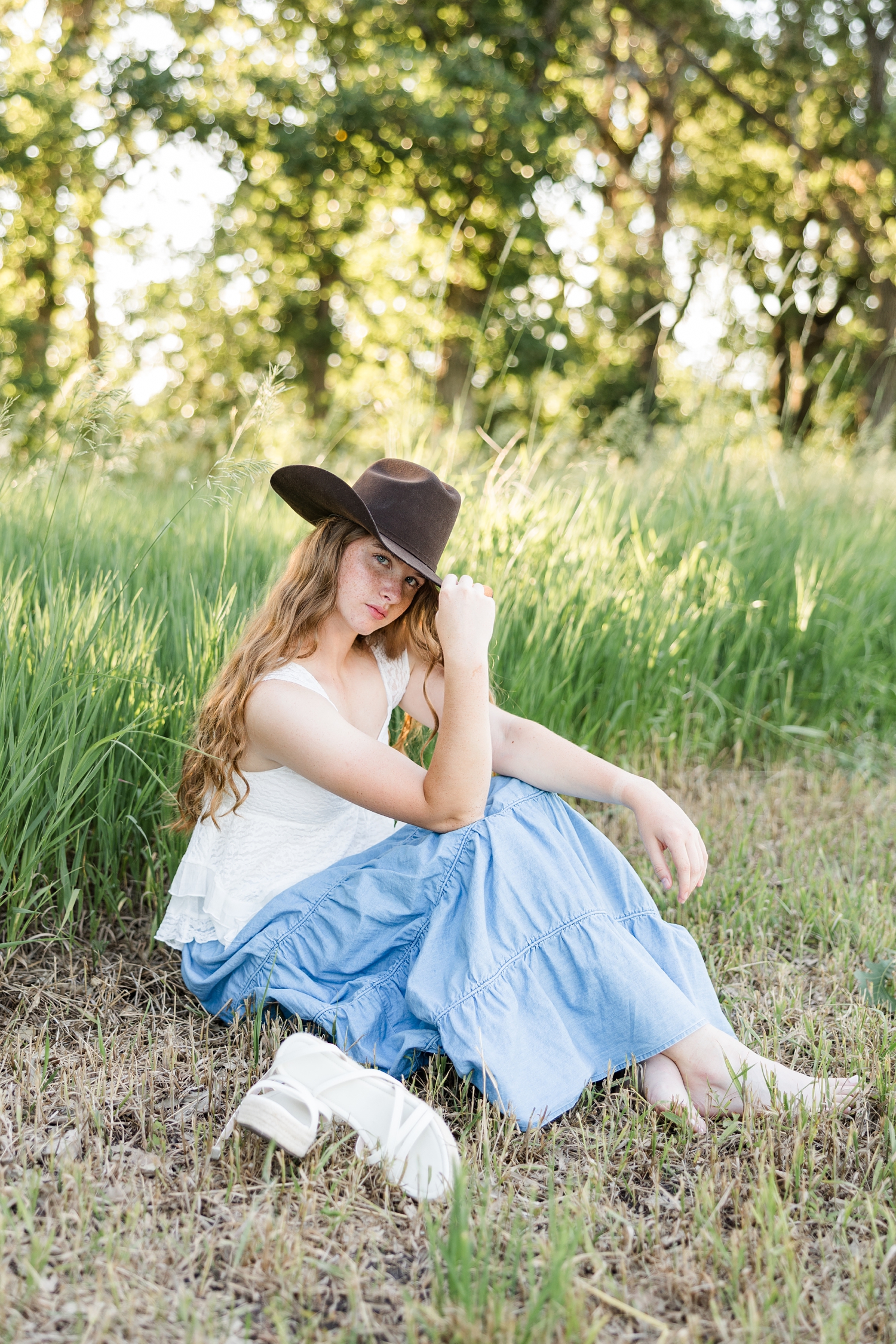 Mya, wearing a white lace top and long, flowy denim skirt and brown cowgirl hat, sits next to tall grass with her knees up and her arms resting on her knees as she tips her hat towards the camera | CB Studio